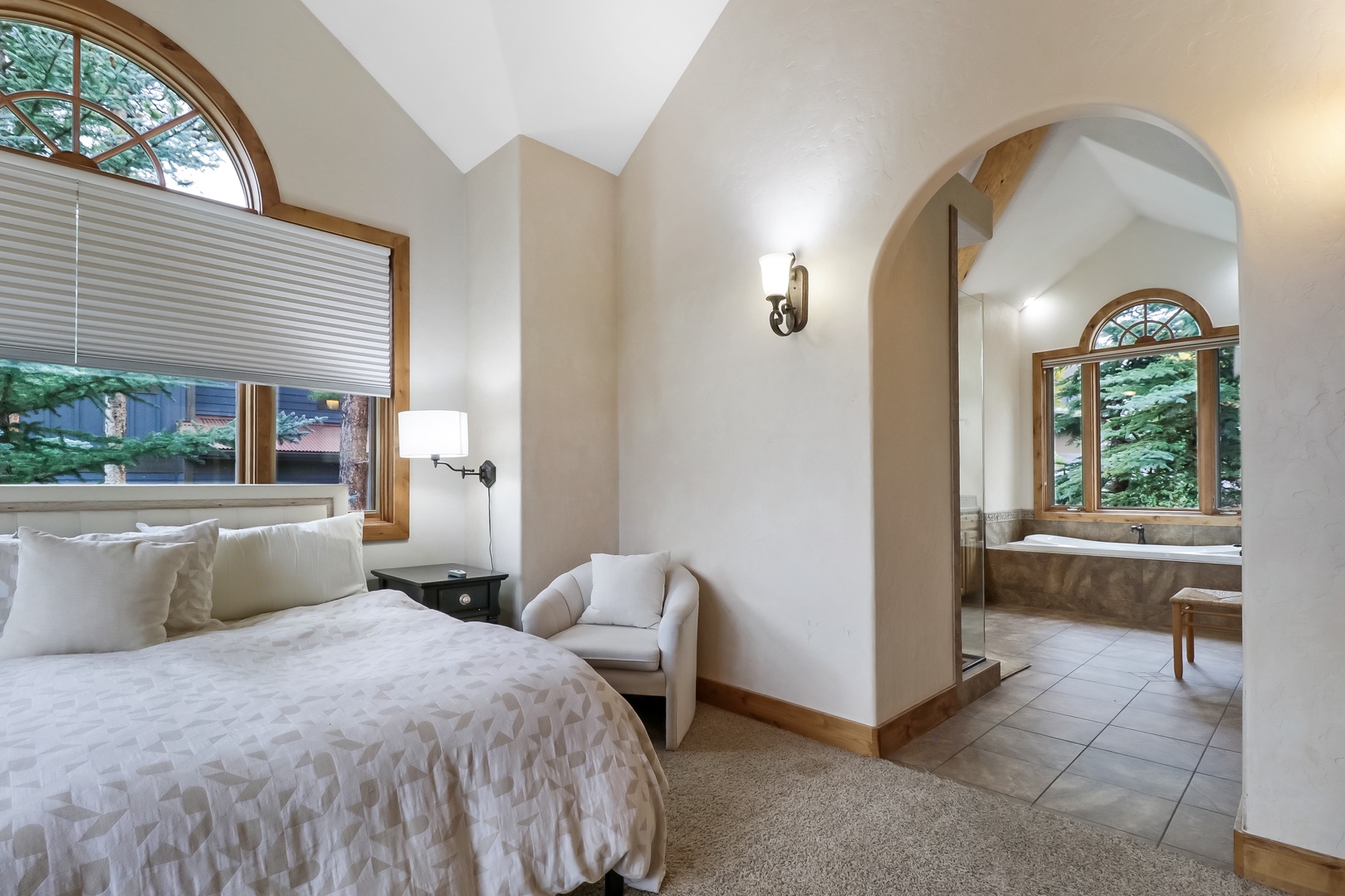 Unwind in your peaceful bedroom with soft linens, vaulted ceilings, and natural light streaming through arched windows. A cozy reading chair invites relaxation, while your private ensuite awaits just beyond the elegant archway.