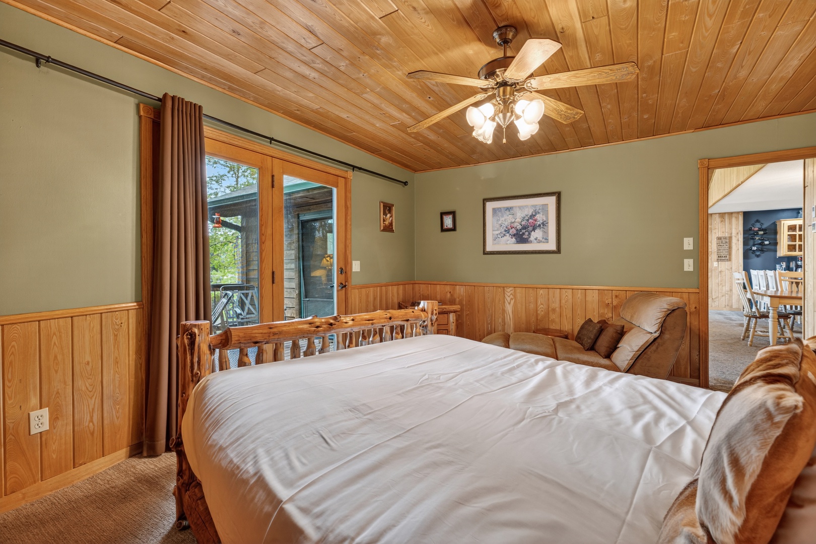 Unwind in this cozy main floor bedroom featuring a plush bed, warm wood paneling, and charming ceiling fan. Large windows invite natural light and views of the surrounding greenery, creating your perfect retreat.