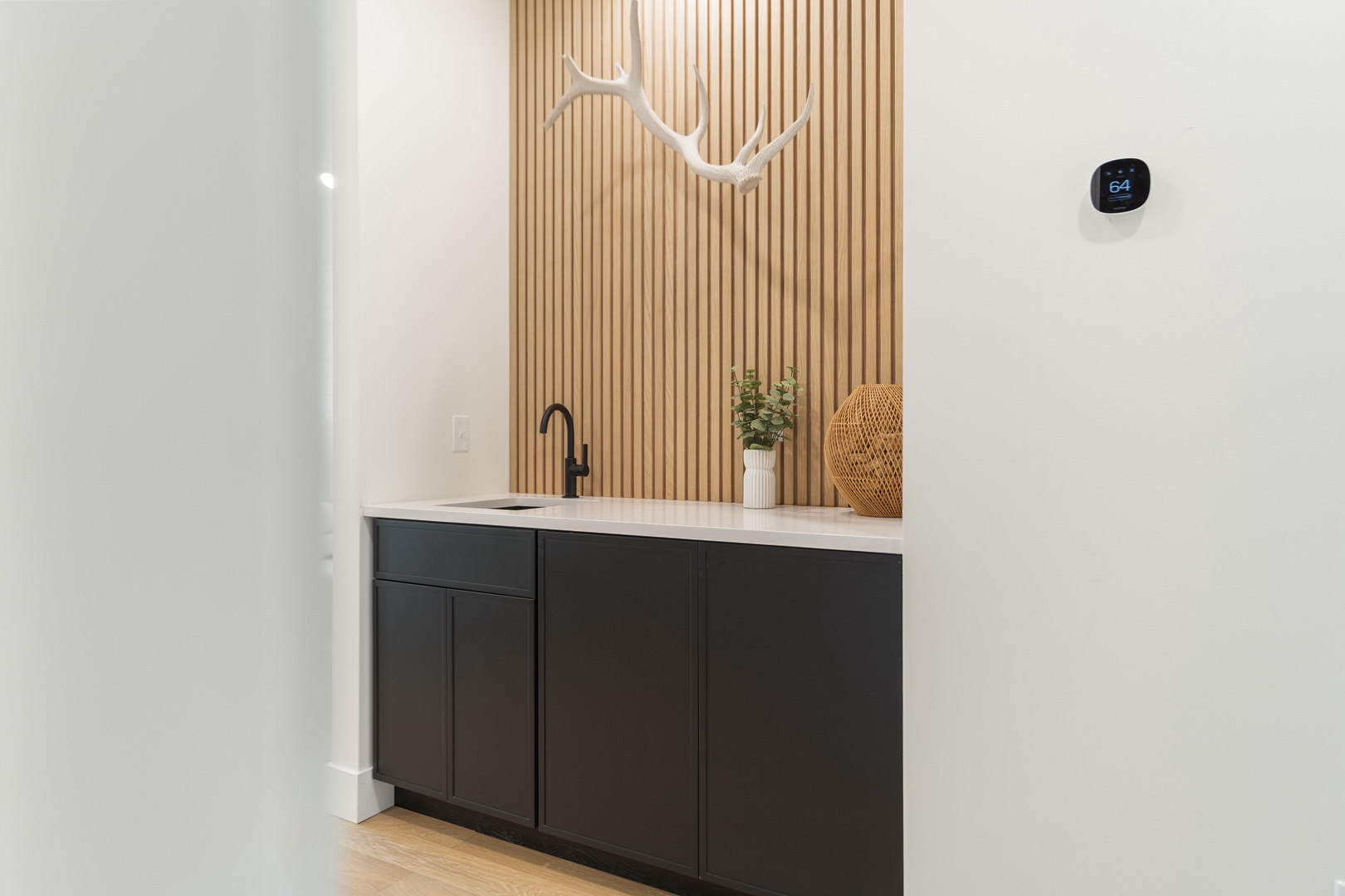 Your stylish wet bar area features sleek dark cabinetry with a white countertop and modern black fixtures, set against a striking wood-slat accent wall with decorative touches.