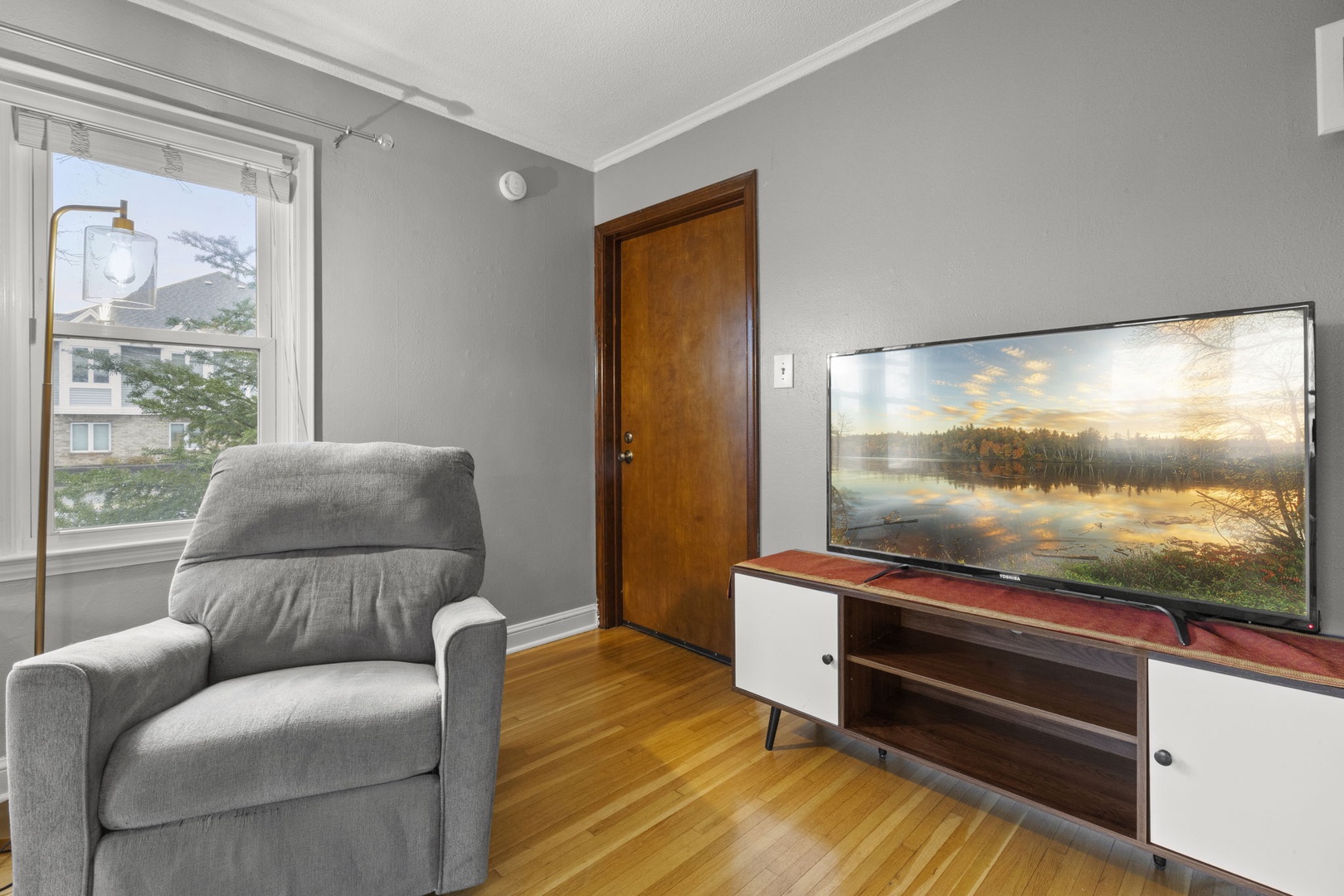 Unwind in your cozy living space with a plush armchair and large screen—perfect for relaxing after exploring the area. Natural light fills the room, inviting you to settle in.