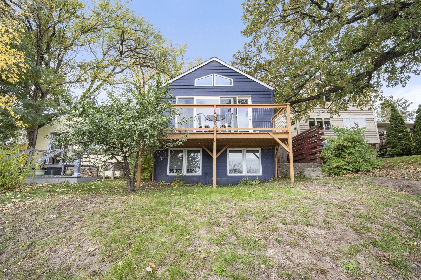 A charming two-story vacation home nestled among mature trees, featuring a spacious elevated deck and surrounded by natural greenery. The property offers a peaceful retreat with outdoor space for relaxation.