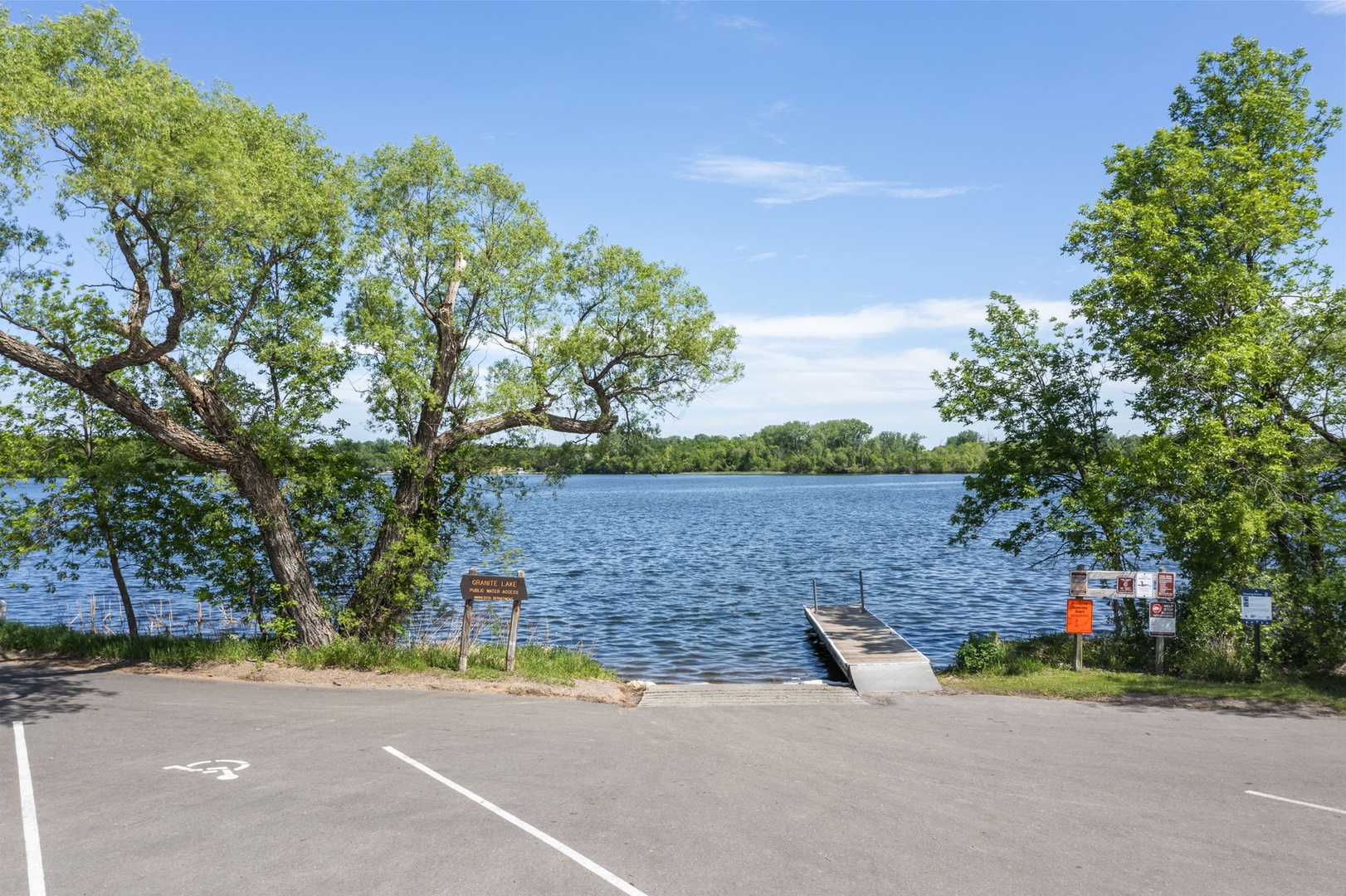 A peaceful lake access with dock and boat launch, framed by mature trees under sunny skies. Convenient parking completes this scenic outdoor escape.