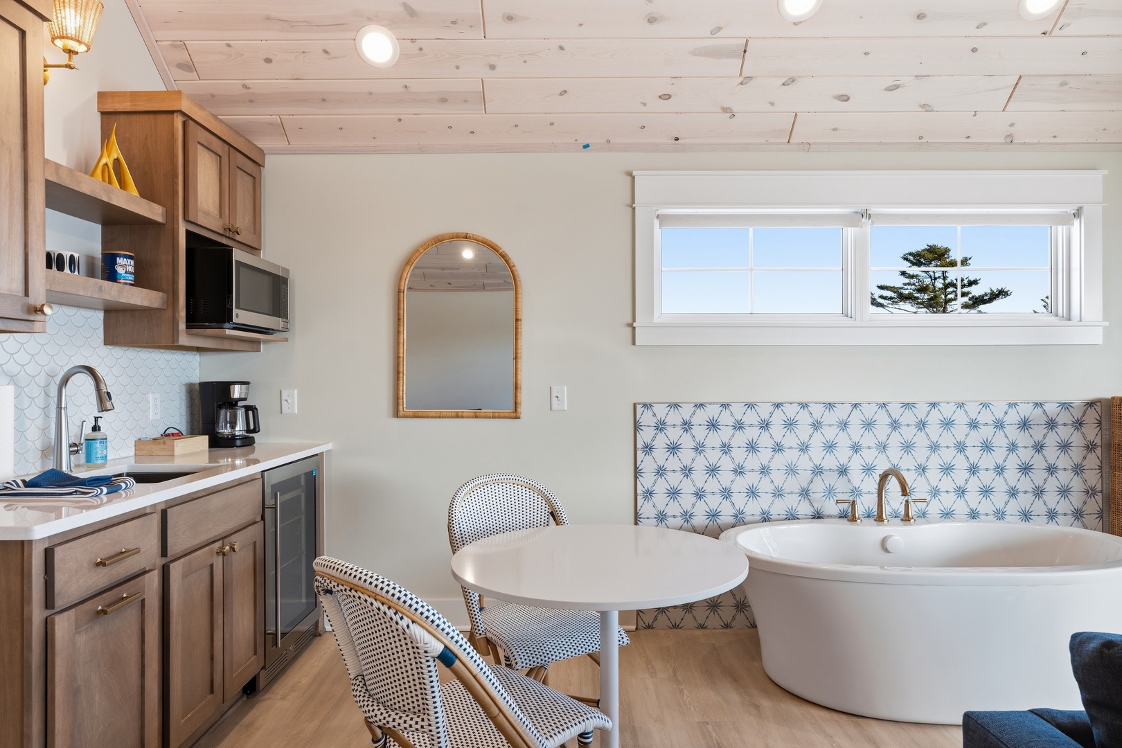 Step into your charming coastal retreat, where a cozy kitchenette and a dreamy soaking tub meet under white pine ceilings, with glimpses of the shoreline through the window.
