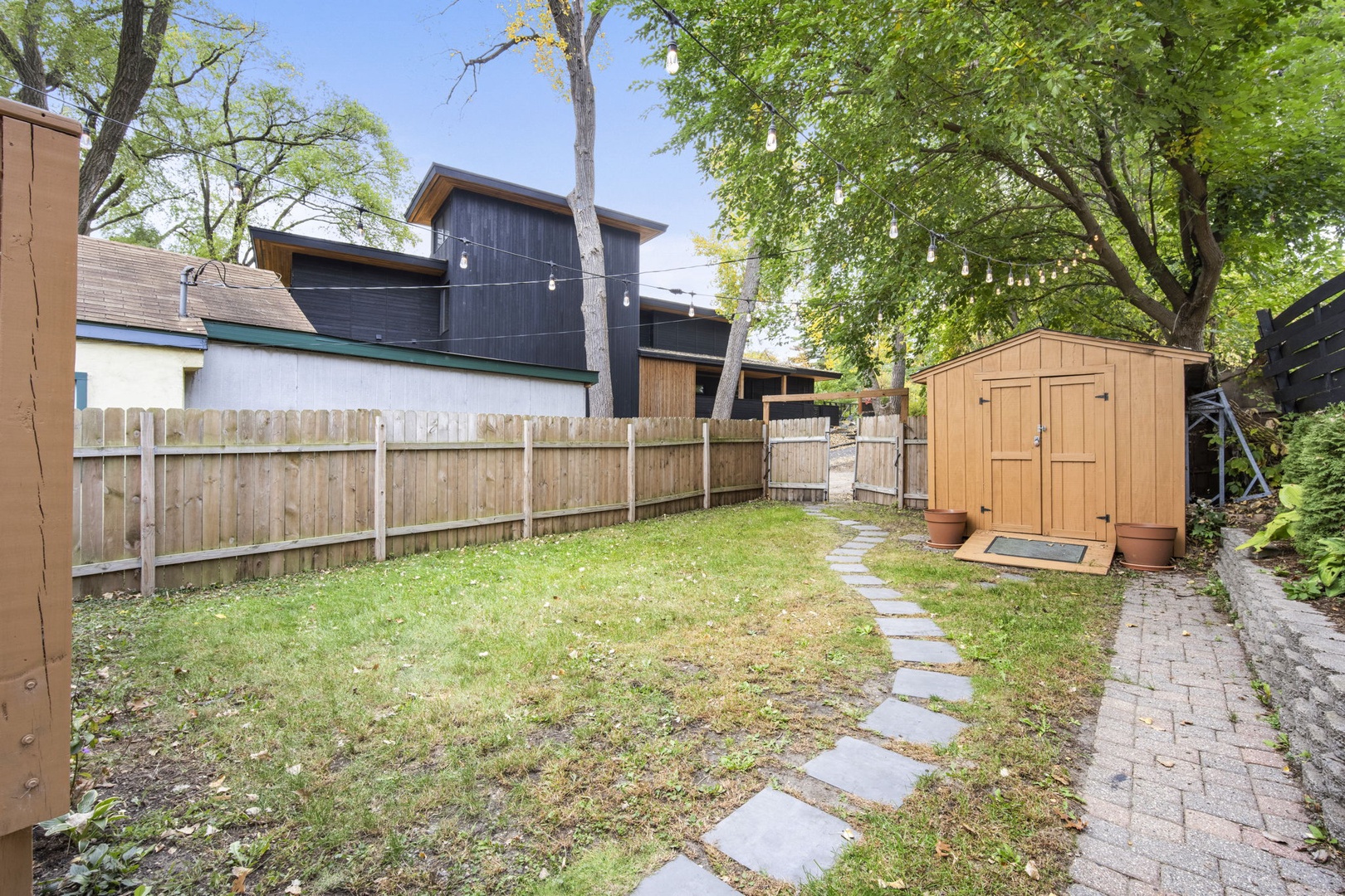 A peaceful backyard featuring a modern property surrounded by mature trees, with a stone pathway, spacious lawn, and garden shed nestled in a quiet neighborhood.