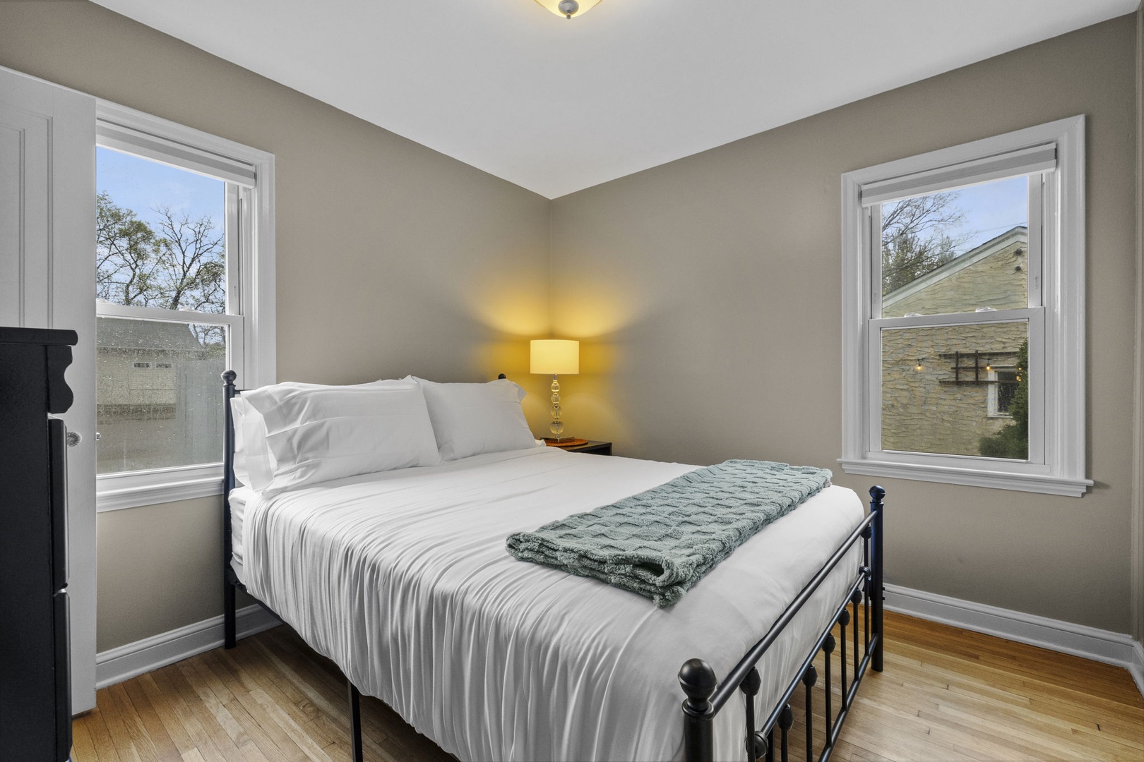 Your peaceful retreat features a plush bed dressed in crisp white linens, flanked by two bright windows that fill the space with natural light and a warm bedside lamp for cozy evenings.
