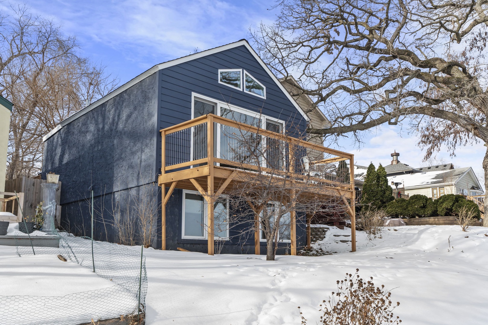 Modern lakeside retreat featuring distinctive blue siding and spacious wooden deck, nestled in a peaceful winter setting with mature trees.