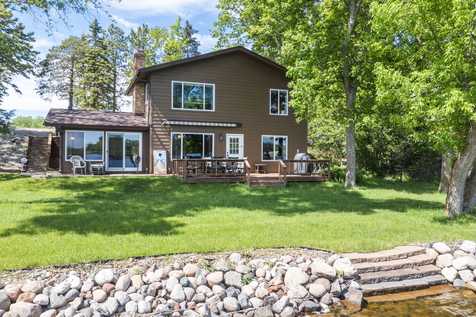 A spacious two-story retreat framed by mature trees and a lush green lawn, with a welcoming deck overlooking waterfront access via a stone-lined pathway and steps to the shore.