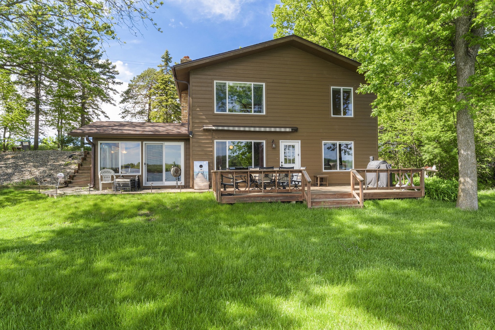 A spacious two-story vacation home nestled among tall trees, featuring a welcoming wooden deck and large windows overlooking manicured lawns, perfect for peaceful getaways surrounded by nature.