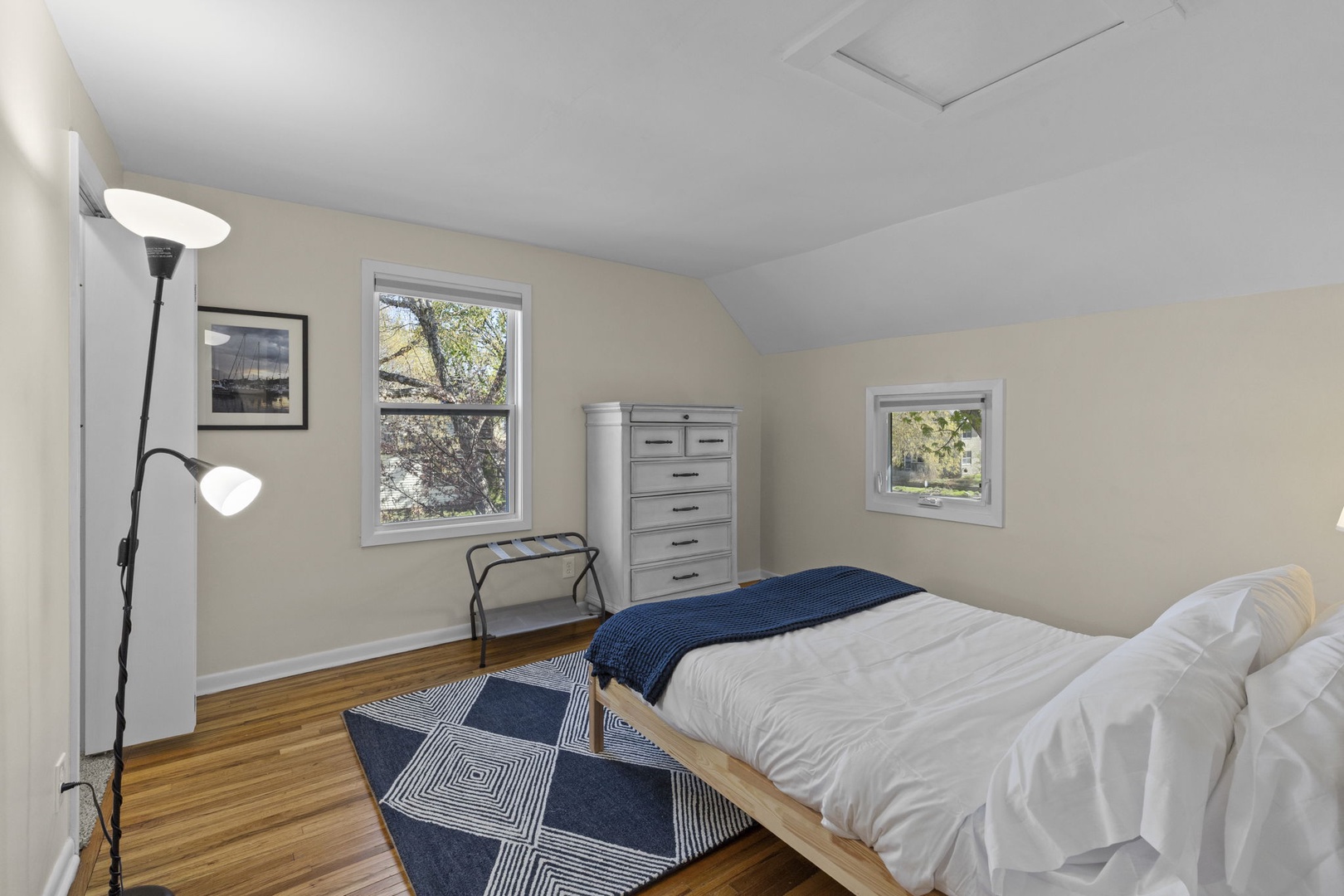 Second bedroom with vaulted ceiling, hardwood floors, white dresser, tall reading lamp, and two windows framing treetop views — light and airy.