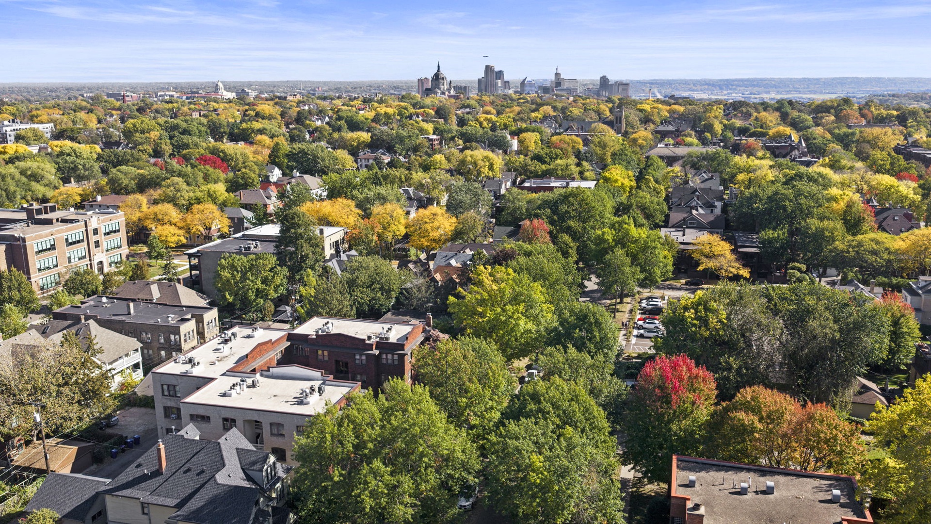 An aerial view capturing the vibrant autumn landscape surrounding the property, with a cityscape visible on the horizon. The neighborhood showcases colorful fall foliage and tree-lined streets.