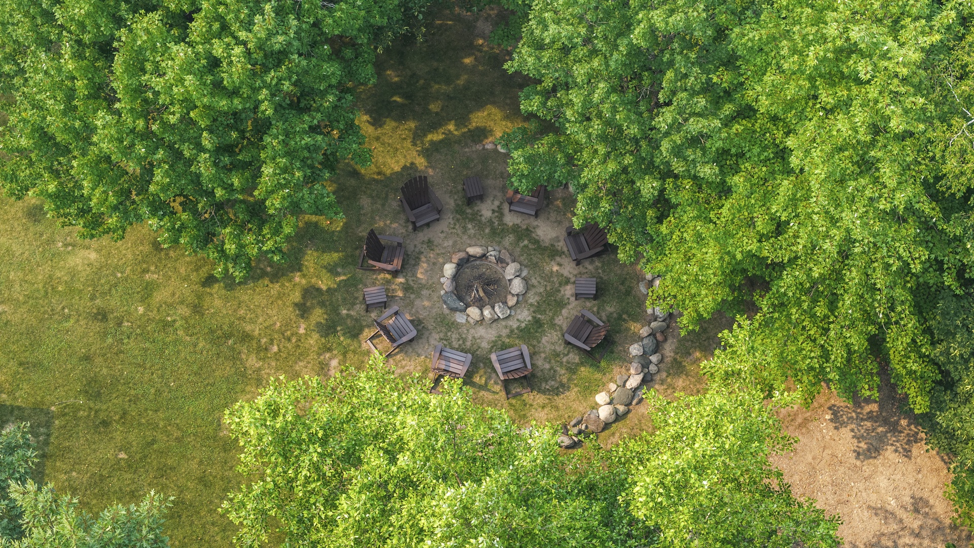 An aerial view reveals the property's secluded outdoor gathering space, nestled within lush forest surroundings. A central stone fire pit anchors the woodland clearing, encircled by Adirondack chairs arranged for cozy evenings under the canopy.