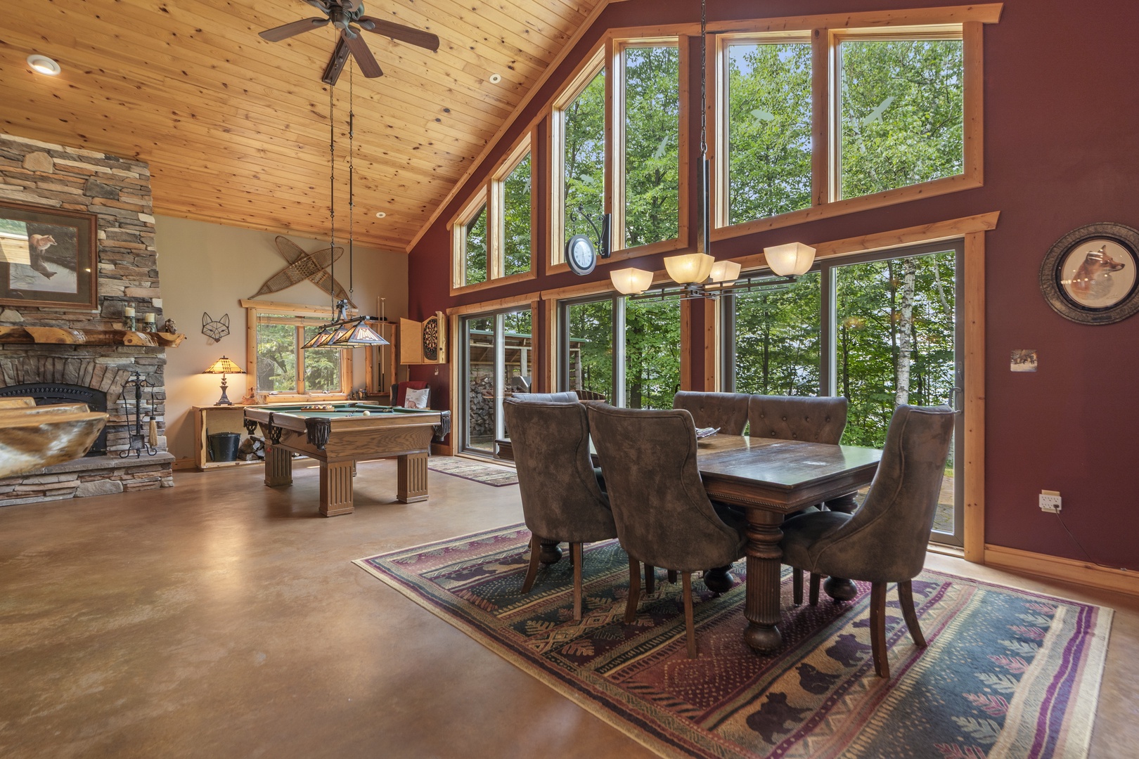 Light pours in through soaring windows overlooking lush greenery, framing your dining and living areas with natural beauty. The vaulted pine ceiling and stone fireplace add warmth to this inviting open space where you can gather and relax.