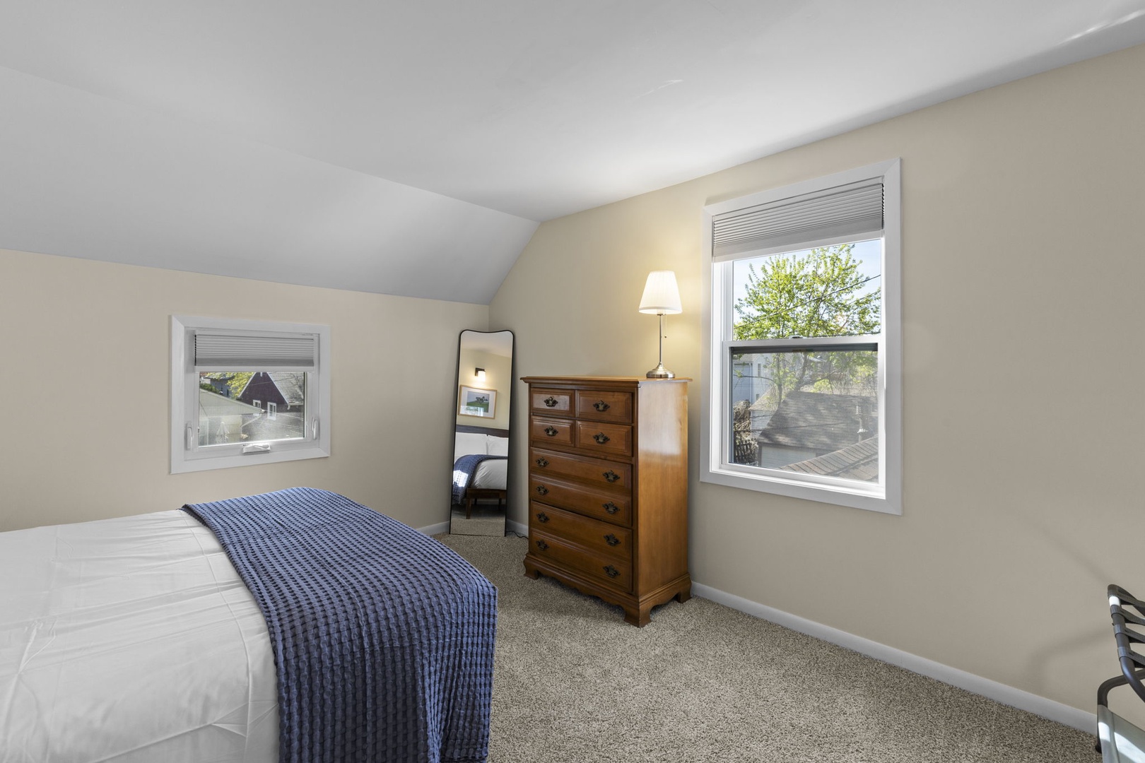 Cozy queen bedroom with vaulted ceiling, plush carpet, tall dresser, full-length mirror, and treetop views — peaceful and private.