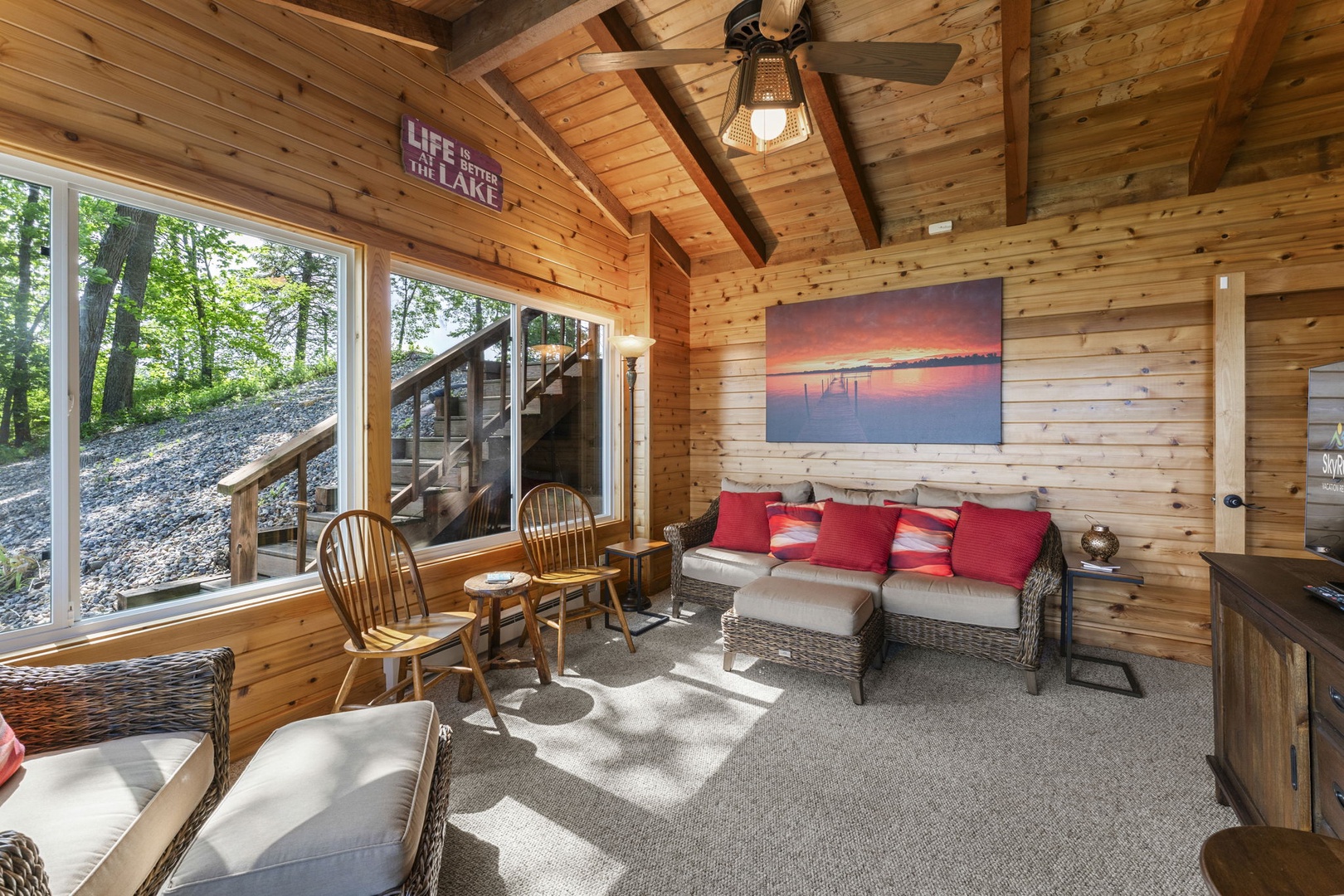 Step into your lakeside retreat—a cozy living room with soaring wood ceilings, wicker seating, and warm cabin charm. Natural light pours in as you unwind near the lake.