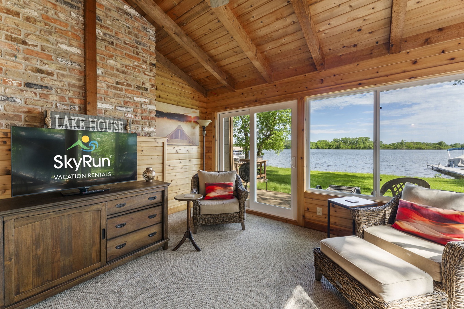 Relax in your sunlit living room with warm wood beams, cozy wicker seating, and sweeping lake views that invite you to unwind and watch the water shimmer just beyond the glass.