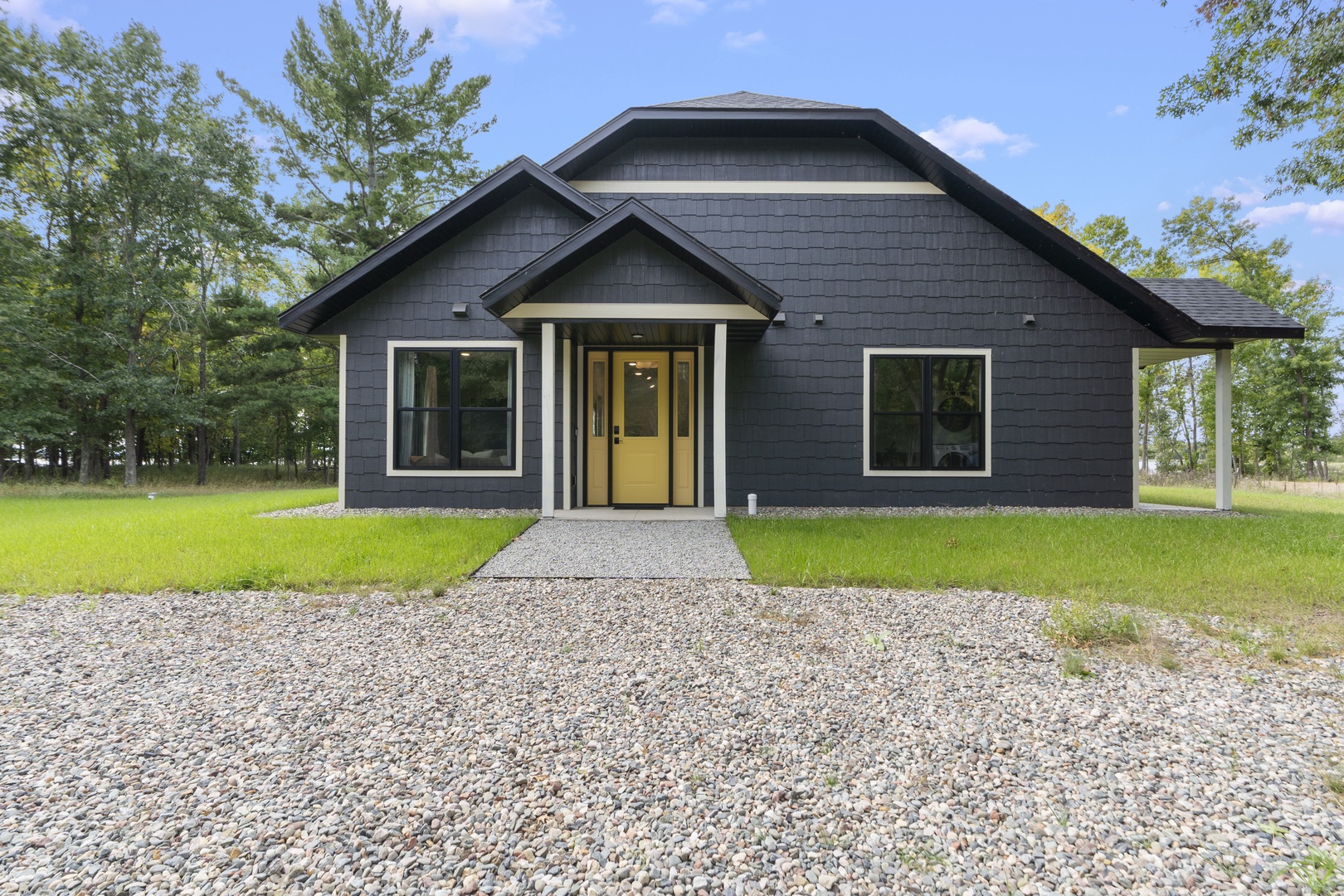 A modern dark-exterior home sits on a peaceful wooded lot with gravel parking and covered porch area.