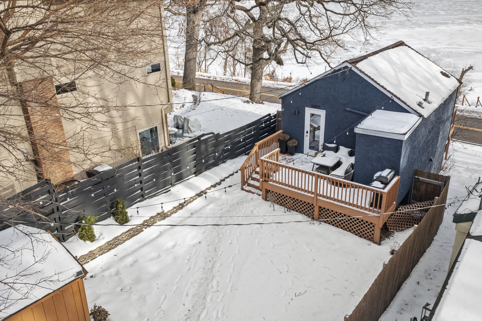 Aerial winter view of property featuring snow-covered outdoor deck and modern blue building surrounded by bare trees and neighboring structures.