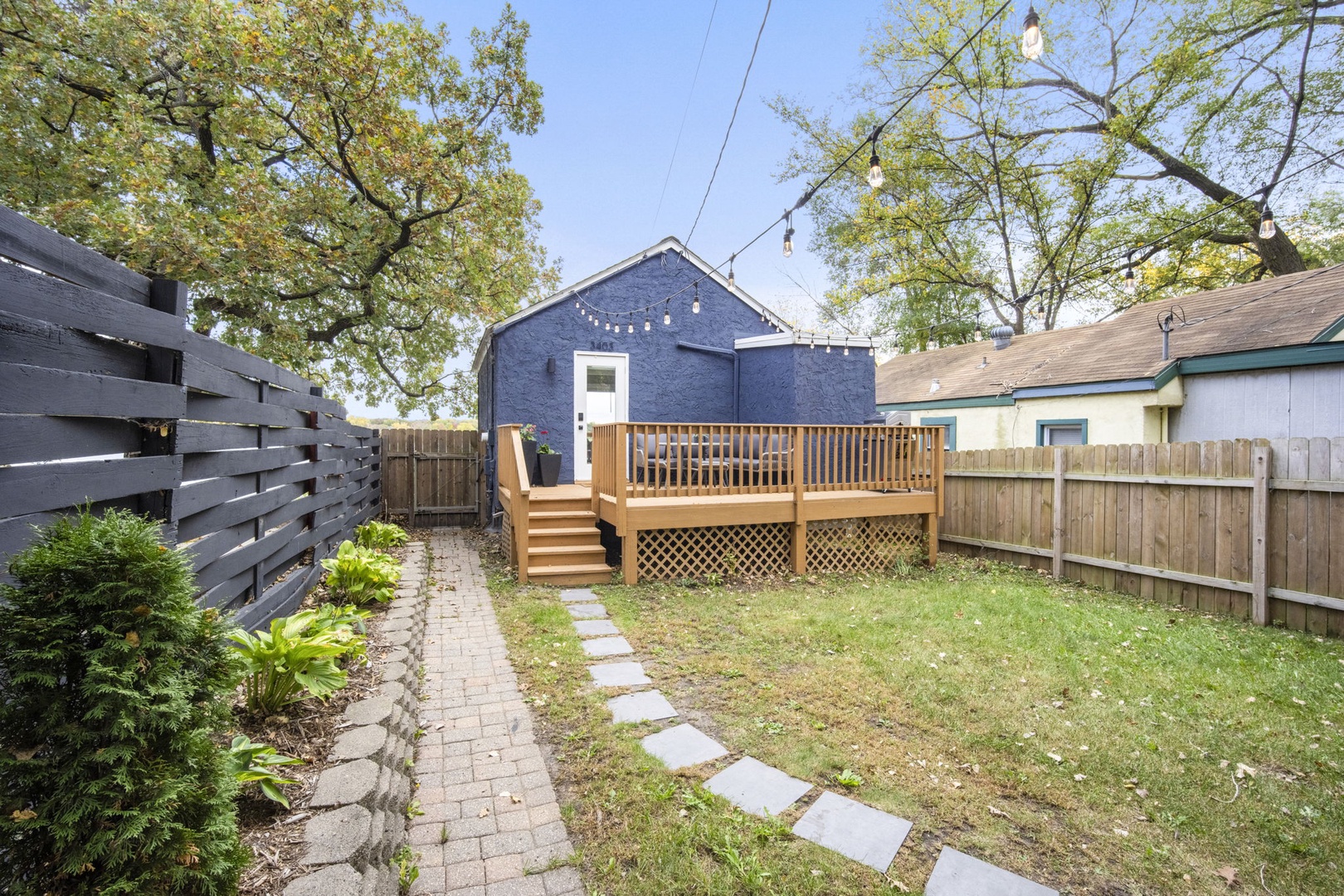 The property features a charming backyard with a raised wood deck, garden pathway, and mature trees providing natural shade and seasonal beauty.
