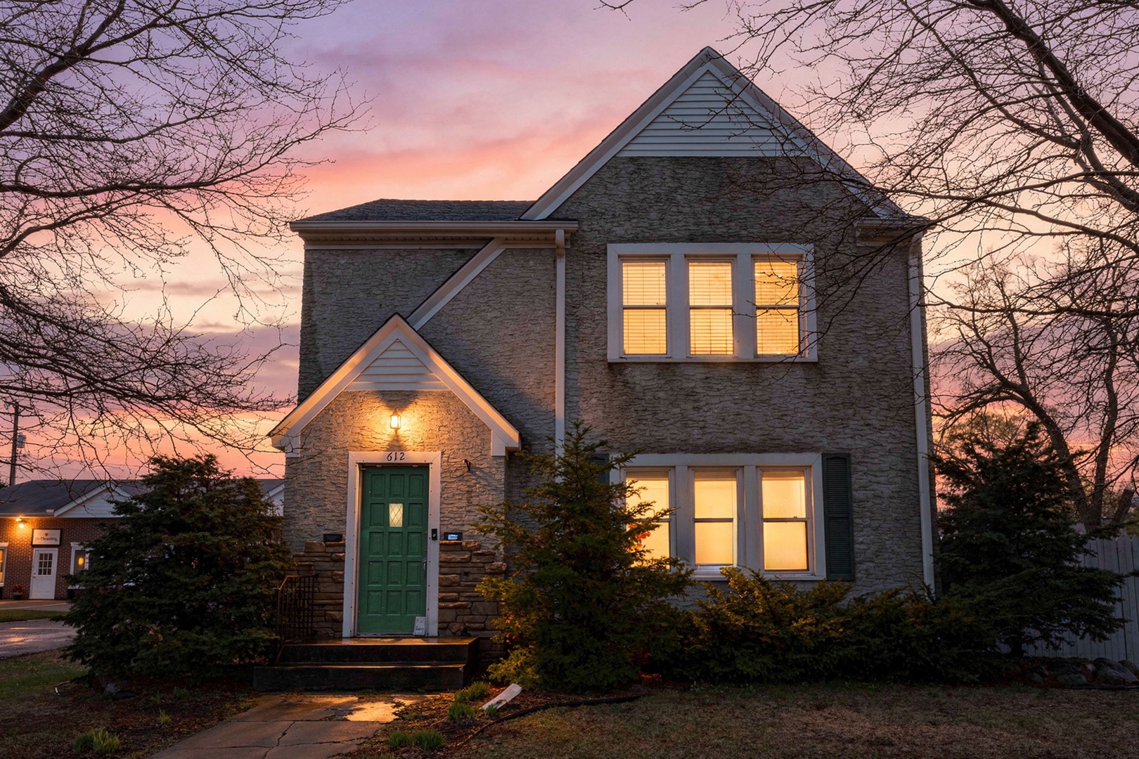 A charming two-story brick home framed by bare trees and evergreens, with warm lights glowing from the windows at dusk and a welcoming green front door marking the entrance.