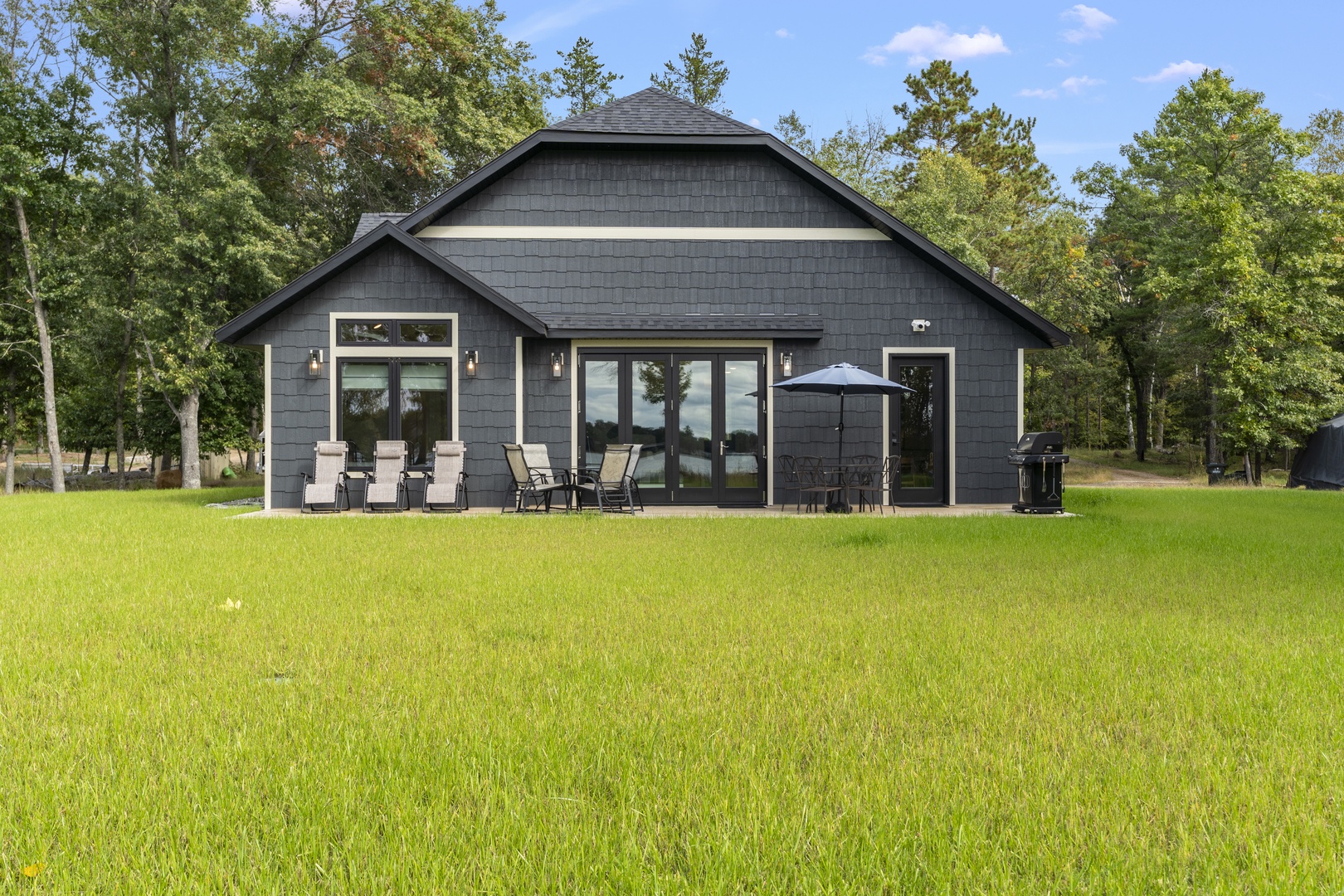 Modern charcoal-grey cottage set on a spacious lawn, surrounded by mature trees. Outdoor seating includes lounge chairs and a covered dining area with grill, providing peaceful natural surroundings for your stay.