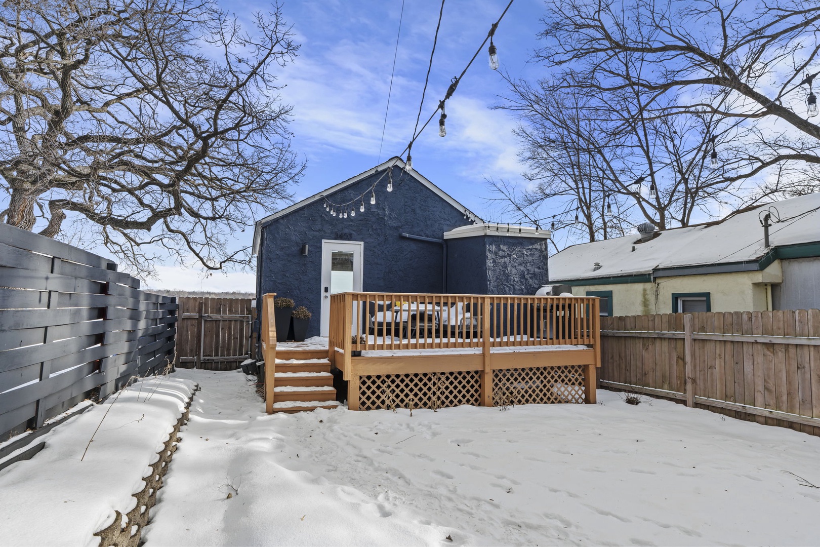 Charming blue cottage with wooden deck surrounded by snow-covered yard and mature trees during winter season.