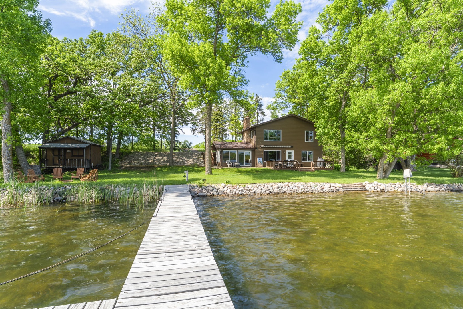 Waterfront property with private dock access, surrounded by mature trees and expansive lawn. The main house features two levels with covered gazebo to the left.