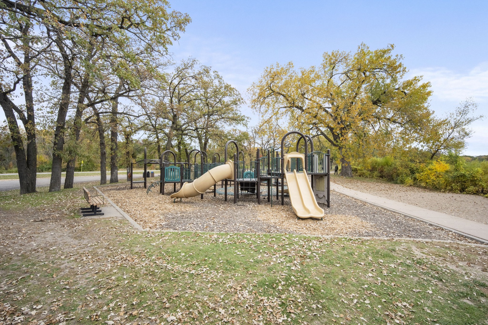 A well-equipped playground featuring slides and climbing structures sits within a peaceful park area surrounded by mature trees with autumn foliage.