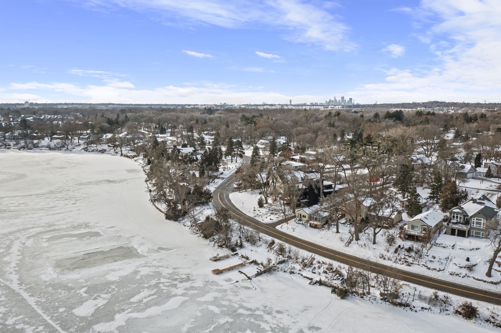 Aerial winter view showcasing the frozen lakefront community with snow-covered homes nestled among mature trees and distant city skyline.