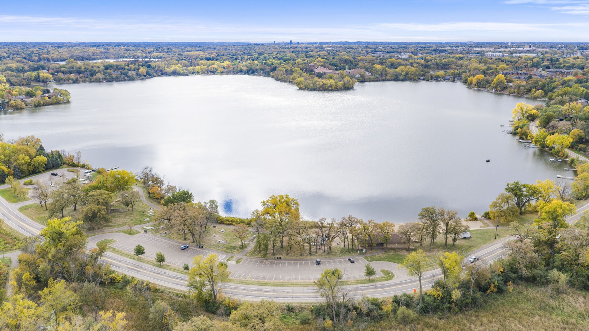An aerial view captures the park's peaceful lakeside location, surrounded by tree-lined shores and open parkland during the autumn season.
