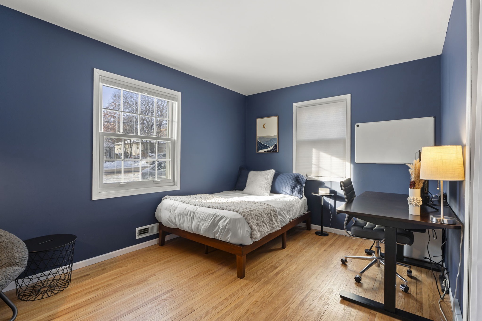 Unwind in your serene blue bedroom with warm hardwood floors, cozy bedding, and a dedicated workspace for productivity.