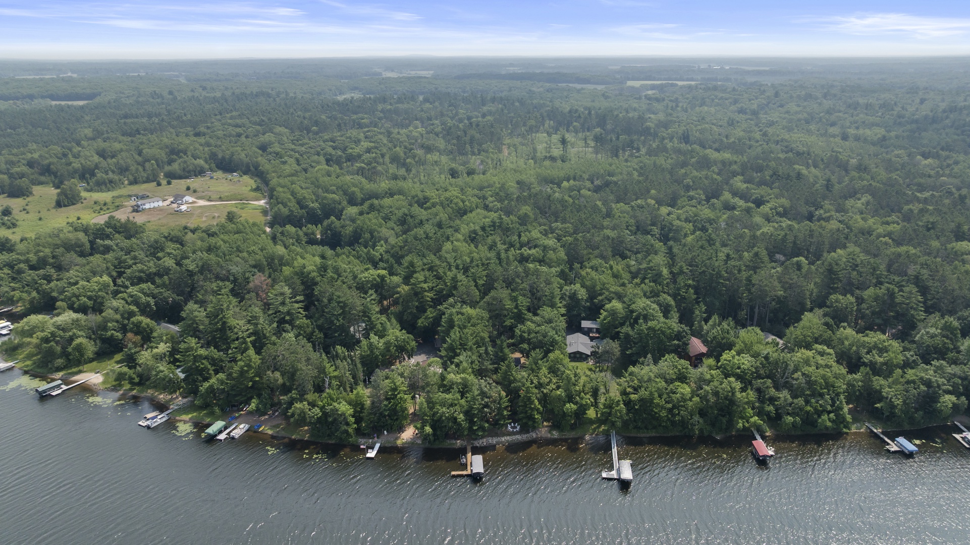 Aerial view reveals a peaceful lakefront property nestled among lush forests, with private docks and boat access. The expansive natural landscape provides a serene retreat surrounded by wooded beauty.