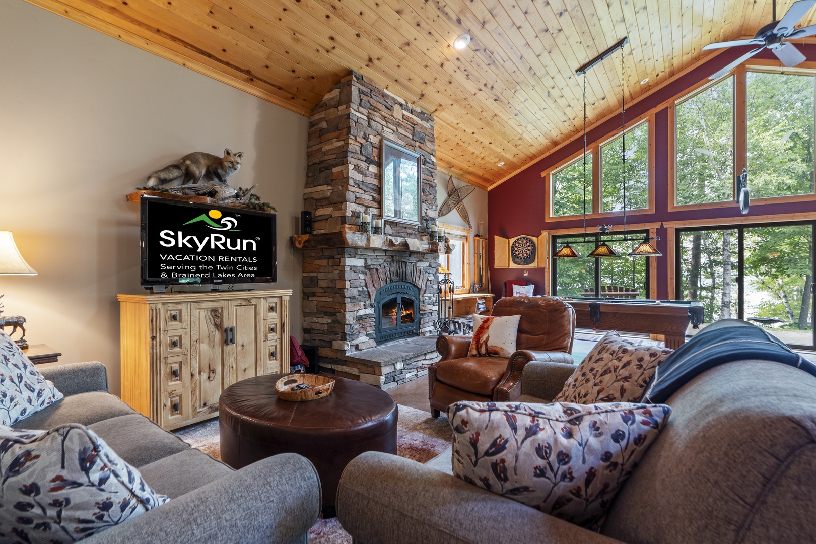 Settle into plush leather seating before the crackling stone fireplace, where soaring pine ceilings and floor-to-ceiling windows frame your peaceful woodland escape.
