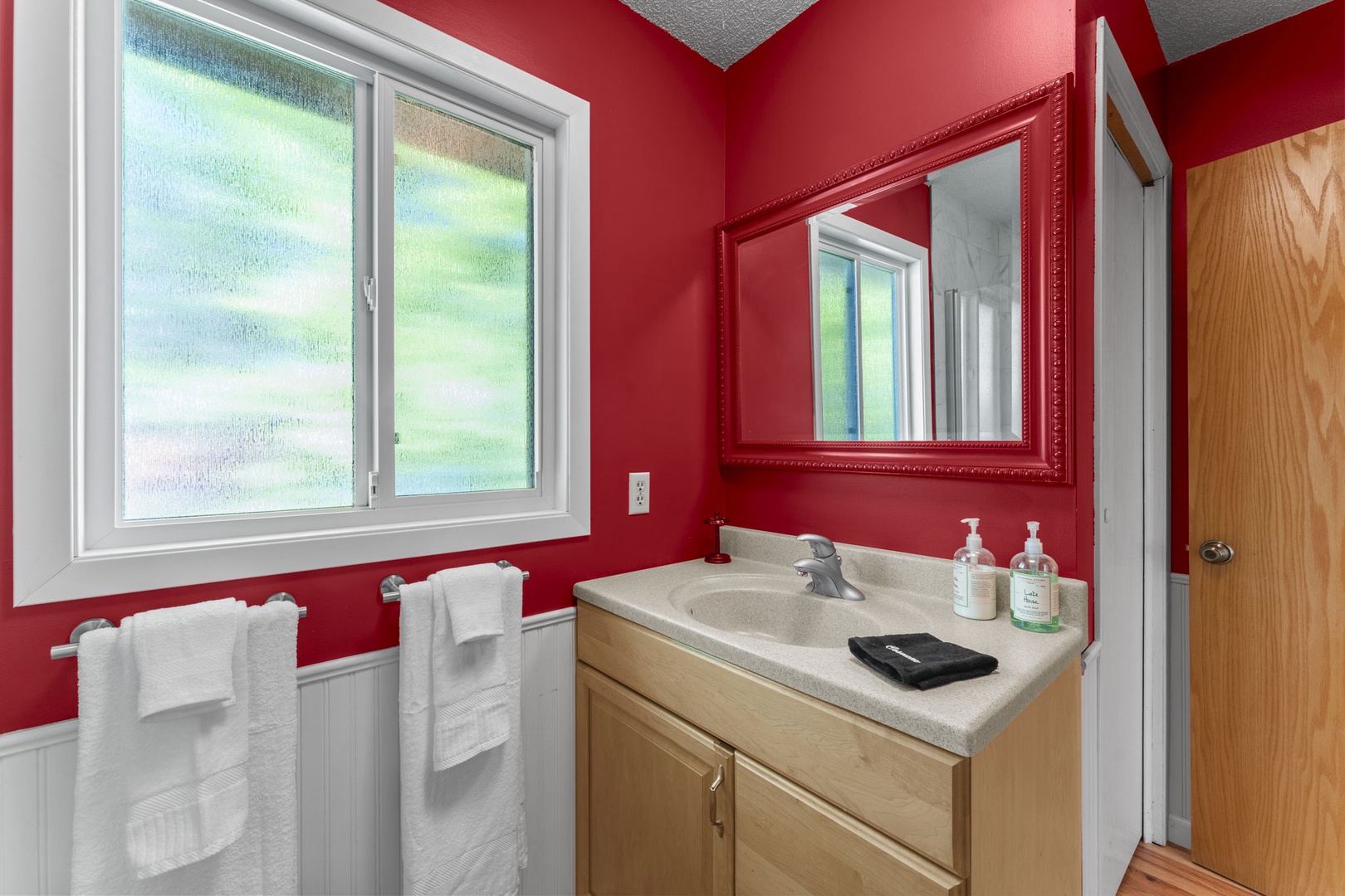 Freshen up in your private bathroom, featuring a spacious vanity, soft towels, and natural light from the window. The bold red walls add a vibrant, welcoming touch to your stay.