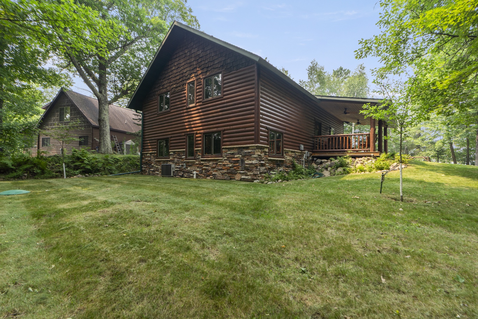 A handsome log cabin nestled among mature trees on a spacious lawn. The property features a covered deck and stone accents, surrounded by lush greenery.