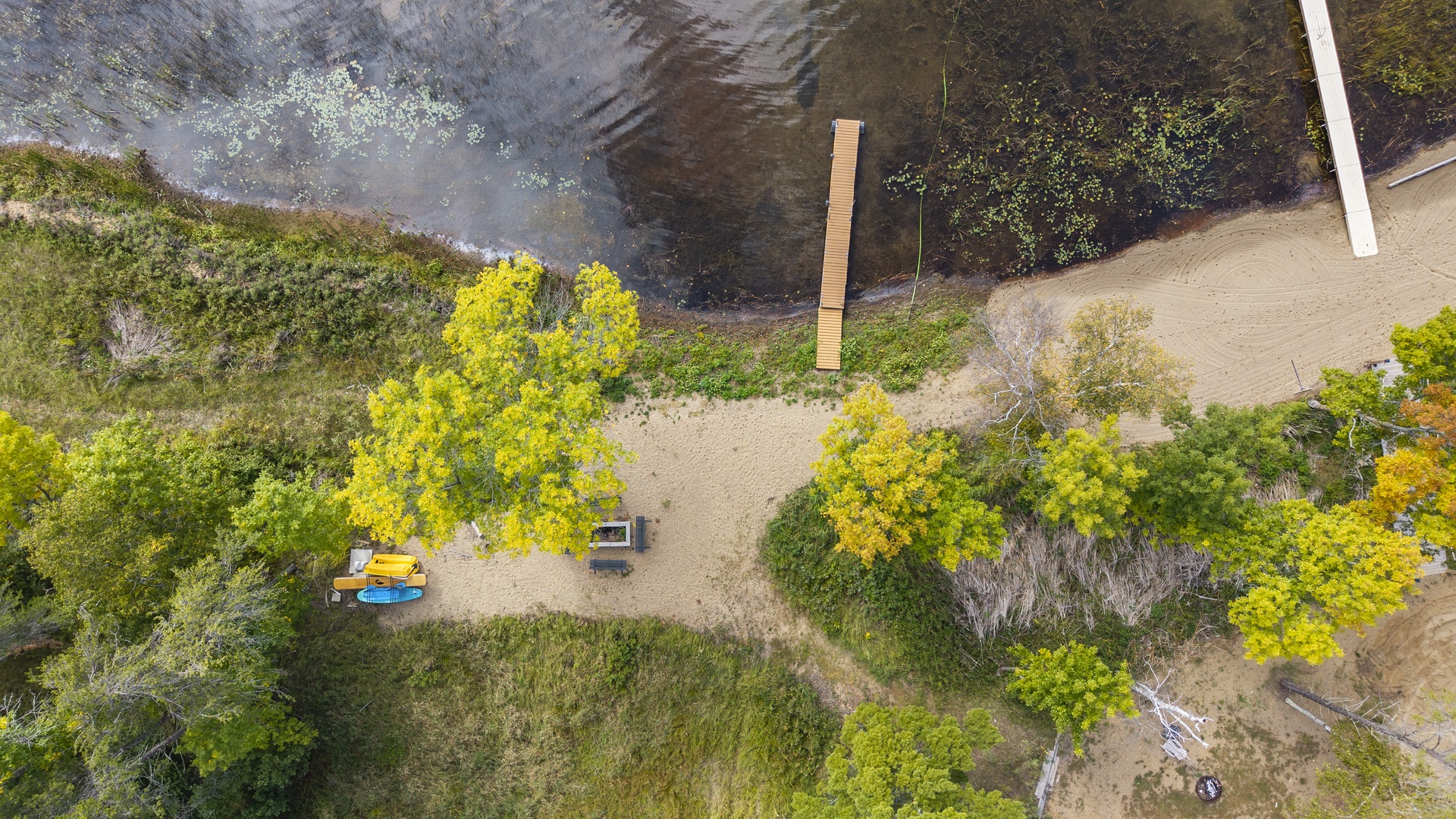 An aerial view showcases the property's serene waterfront setting, surrounded by lush greenery, natural vegetation, and direct water access along a peaceful shoreline. The seasonal foliage and private dock complete the tranquil scene.
