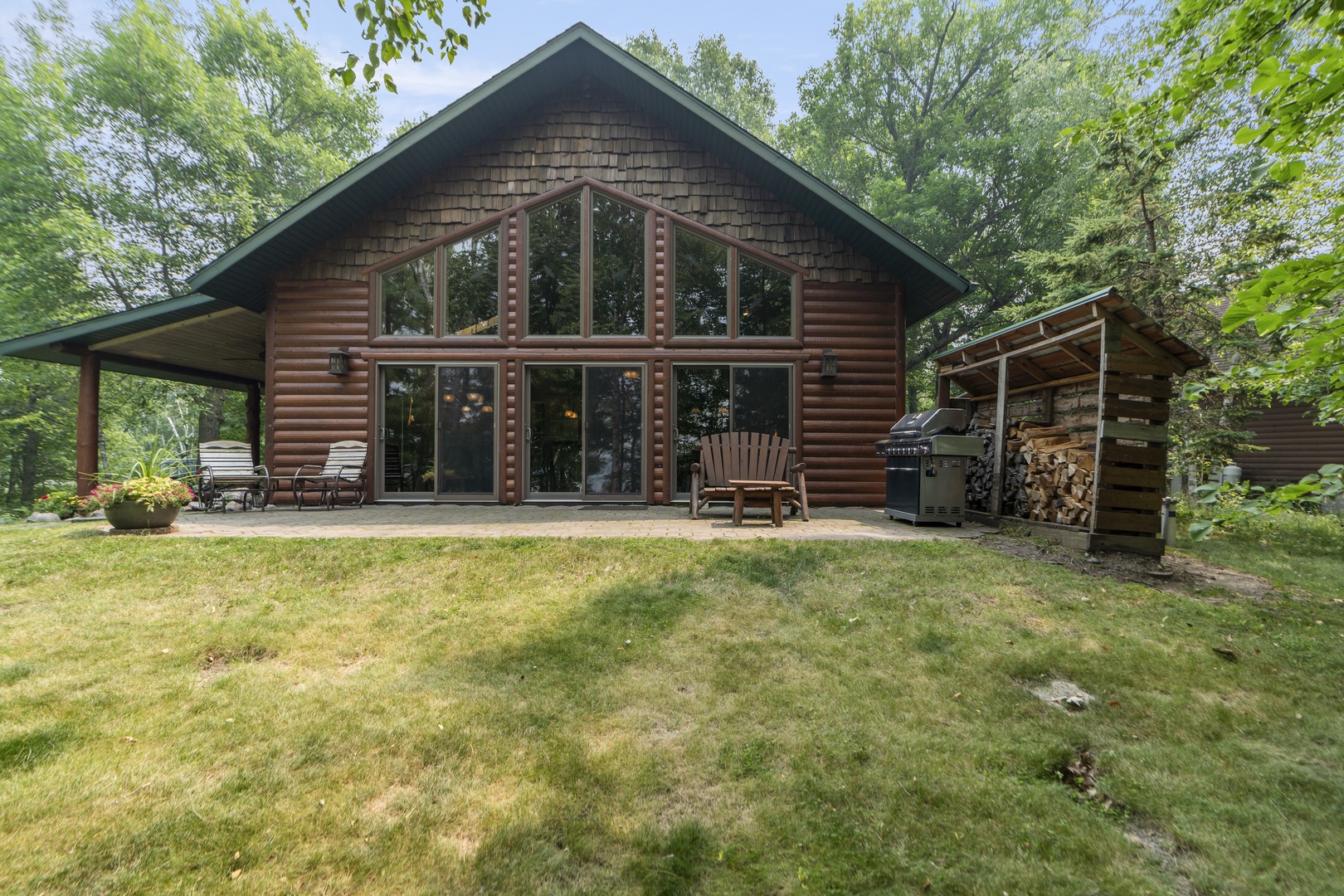 A charming log cabin nestled among mature trees, featuring a covered patio with seating and a BBQ area, all surrounded by a spacious lawn for outdoor gatherings.
