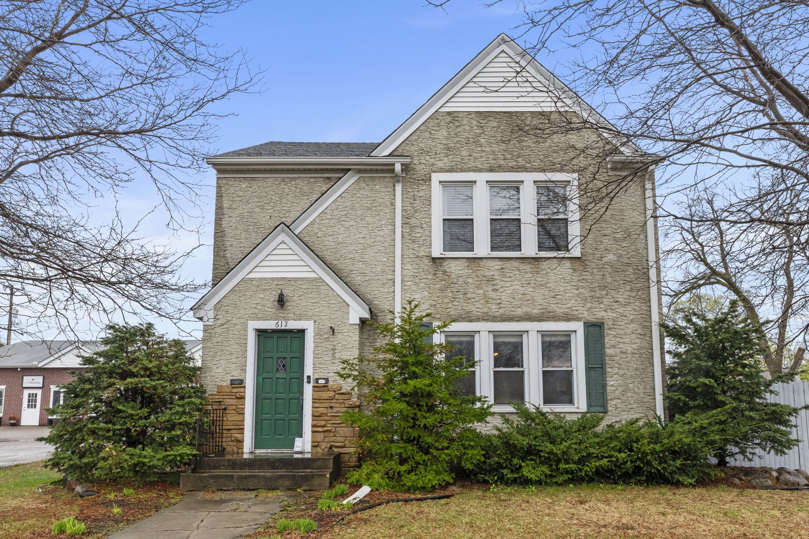 A charming two-story brick property with classic white trim and a welcoming green front door, nestled among mature trees in a quiet residential neighborhood.