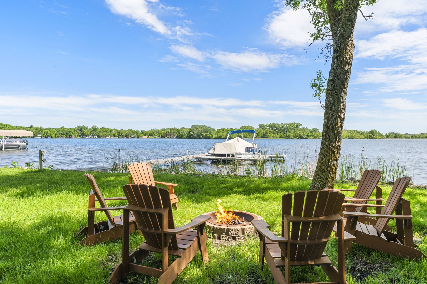 Private lakefront setting with Adirondack chairs around a fire pit. The property includes a boat dock on calm waters, surrounded by lush trees and natural shoreline.