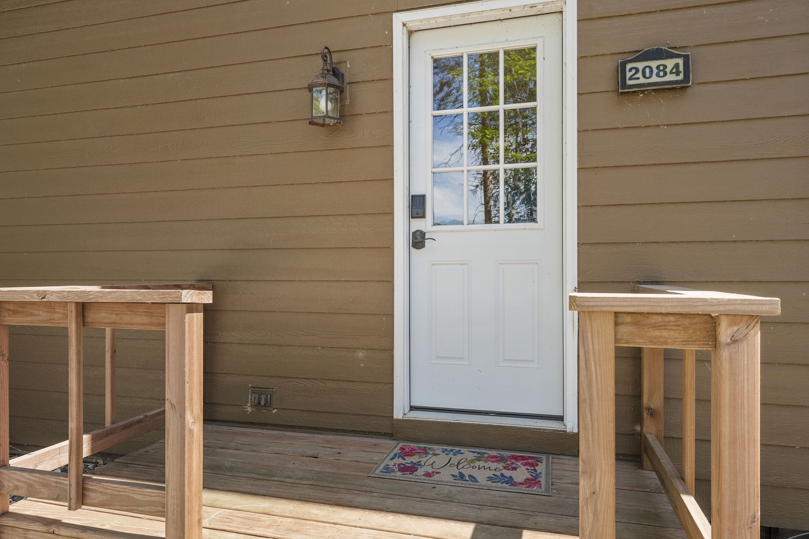 The charming entrance to The Granite Cabin welcomes arrivals with a fresh white door, lantern lighting, and a cheerful floral doormat on a newly constructed wooden deck.