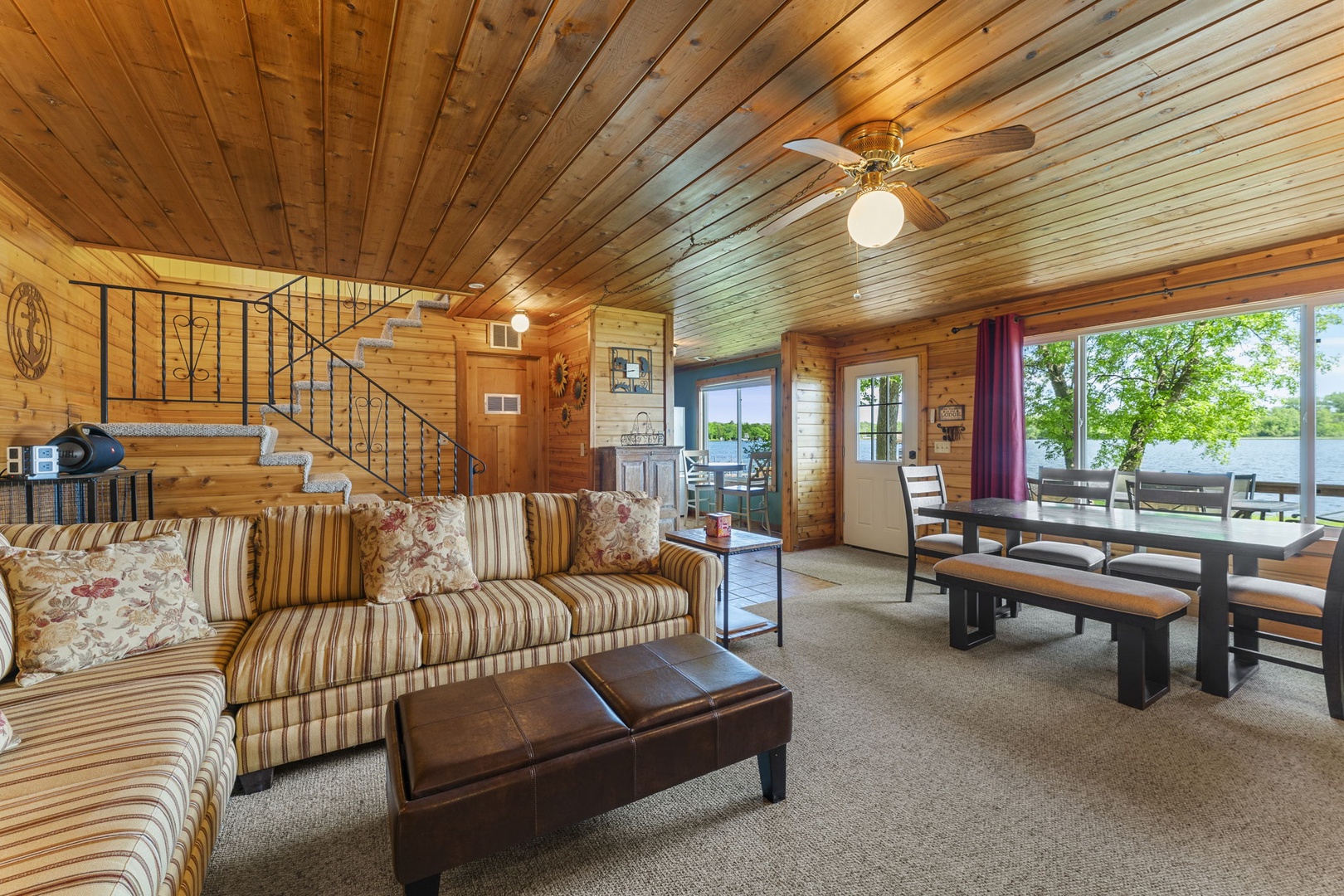 Step into your cozy lakeside retreat with warm wood-planked ceilings and walls that wrap you in rustic comfort. Relax on the plush striped sofa or gather around the handcrafted dining table, where natural light pours through lakefront windows.