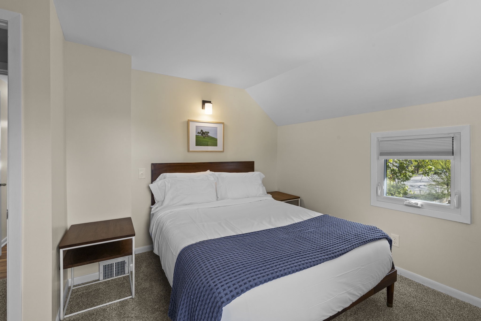 Queen bed with walnut headboard, hotel-quality white linens, wall sconce, and framed art — a serene retreat with soft carpet and garden views.