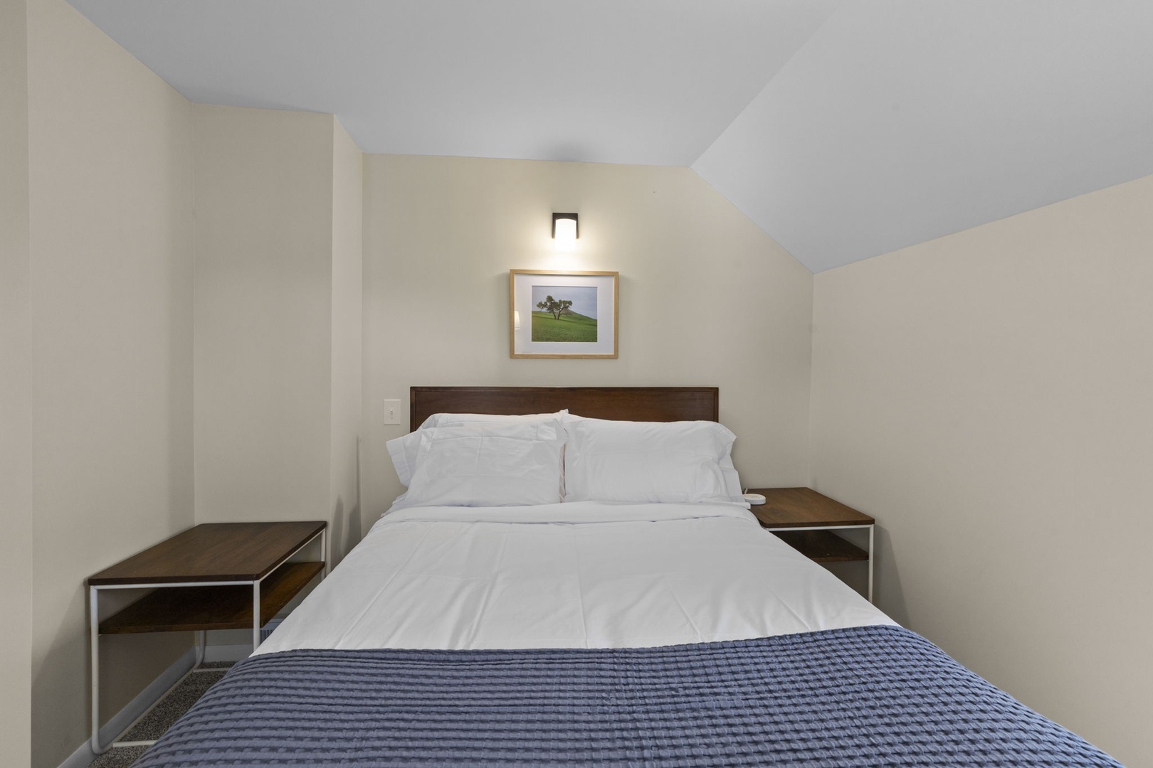 Queen bed close-up with crisp white hotel-style bedding, flanking nightstands, and warm wall sconce — rest easy in this restful upstairs bedroom.
