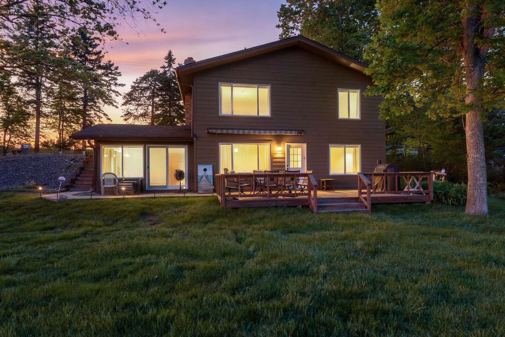 A peaceful lakeside retreat framed by tall pines, glowing warmly as twilight settles over the property. The spacious deck invites evening gatherings beneath a painted sky.