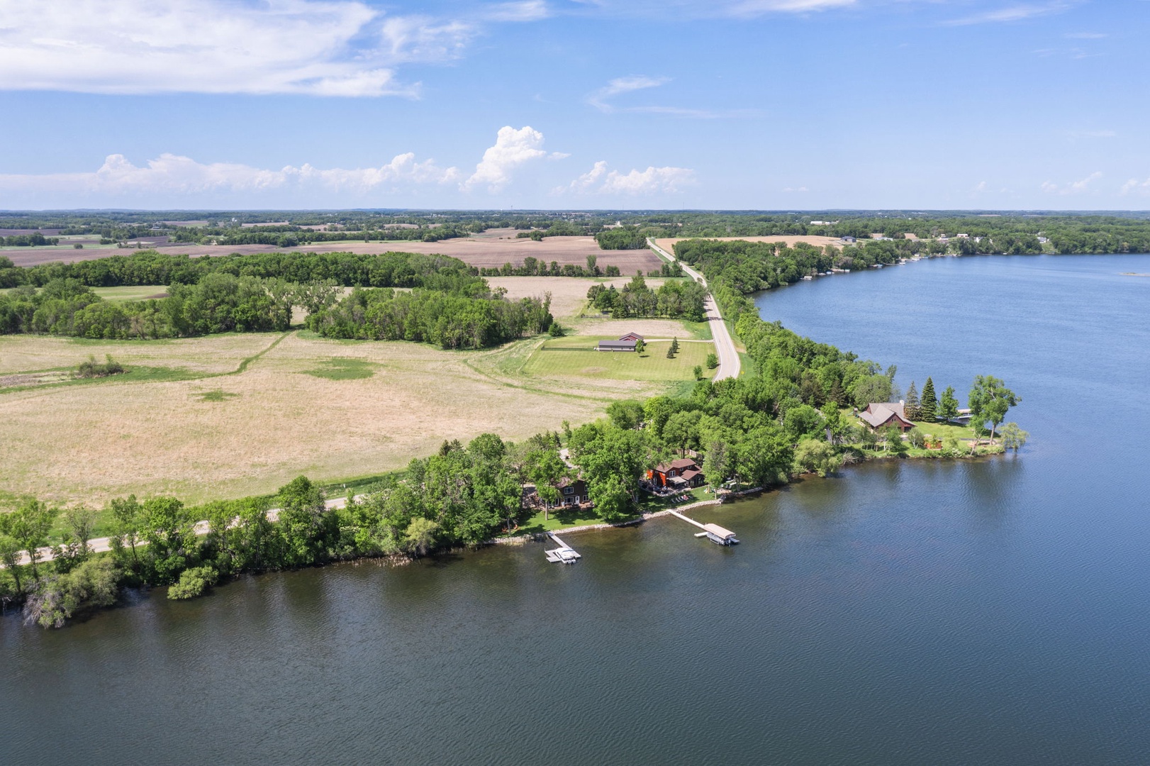 Aerial view of a waterfront vacation rental property nestled along a serene lake, surrounded by lush green landscapes, open fields, and quiet countryside roads.
