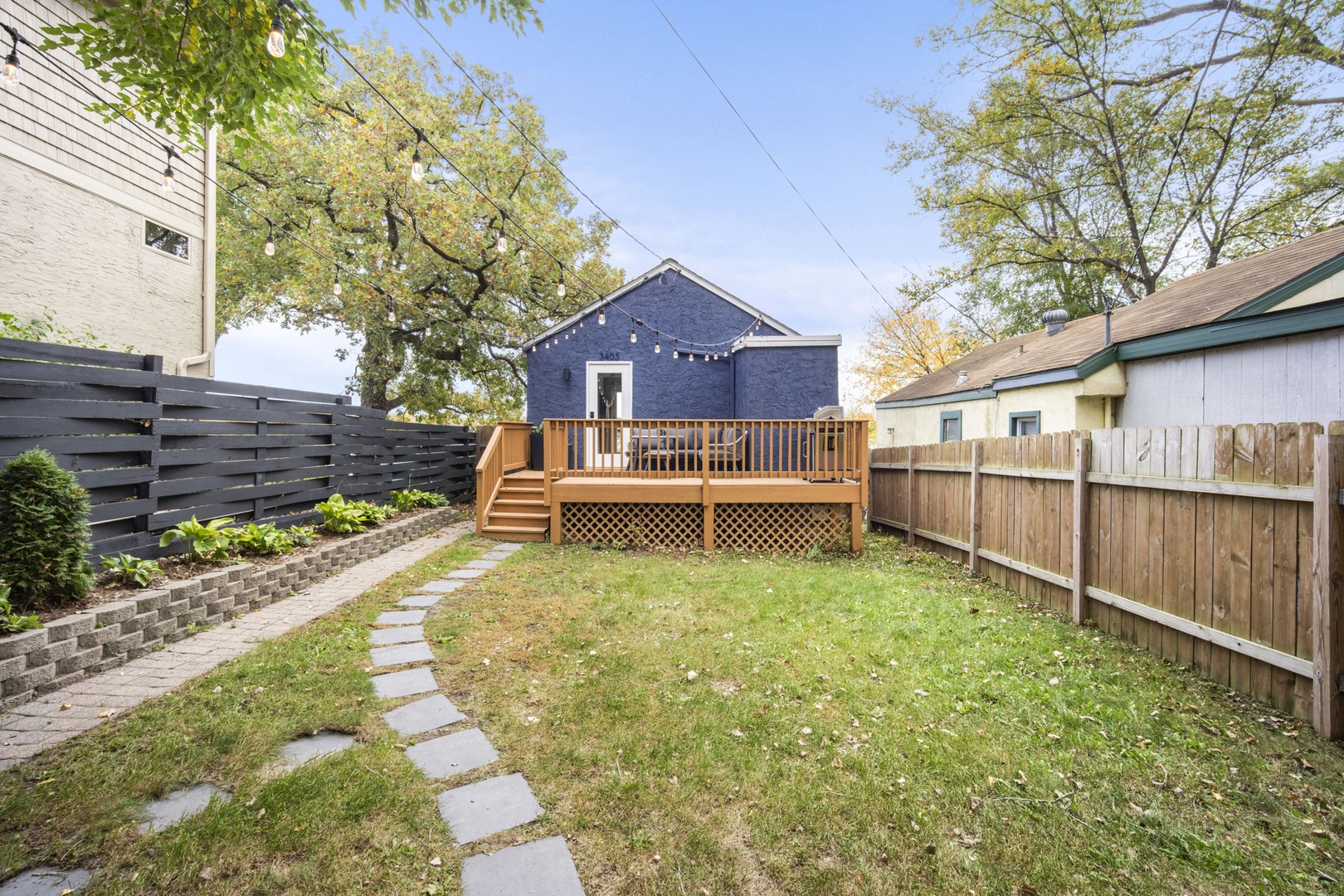 This property features a spacious backyard with a raised wooden deck, fenced-in lawn, and stone walkway leading through a landscaped garden area, offering a peaceful outdoor setting.