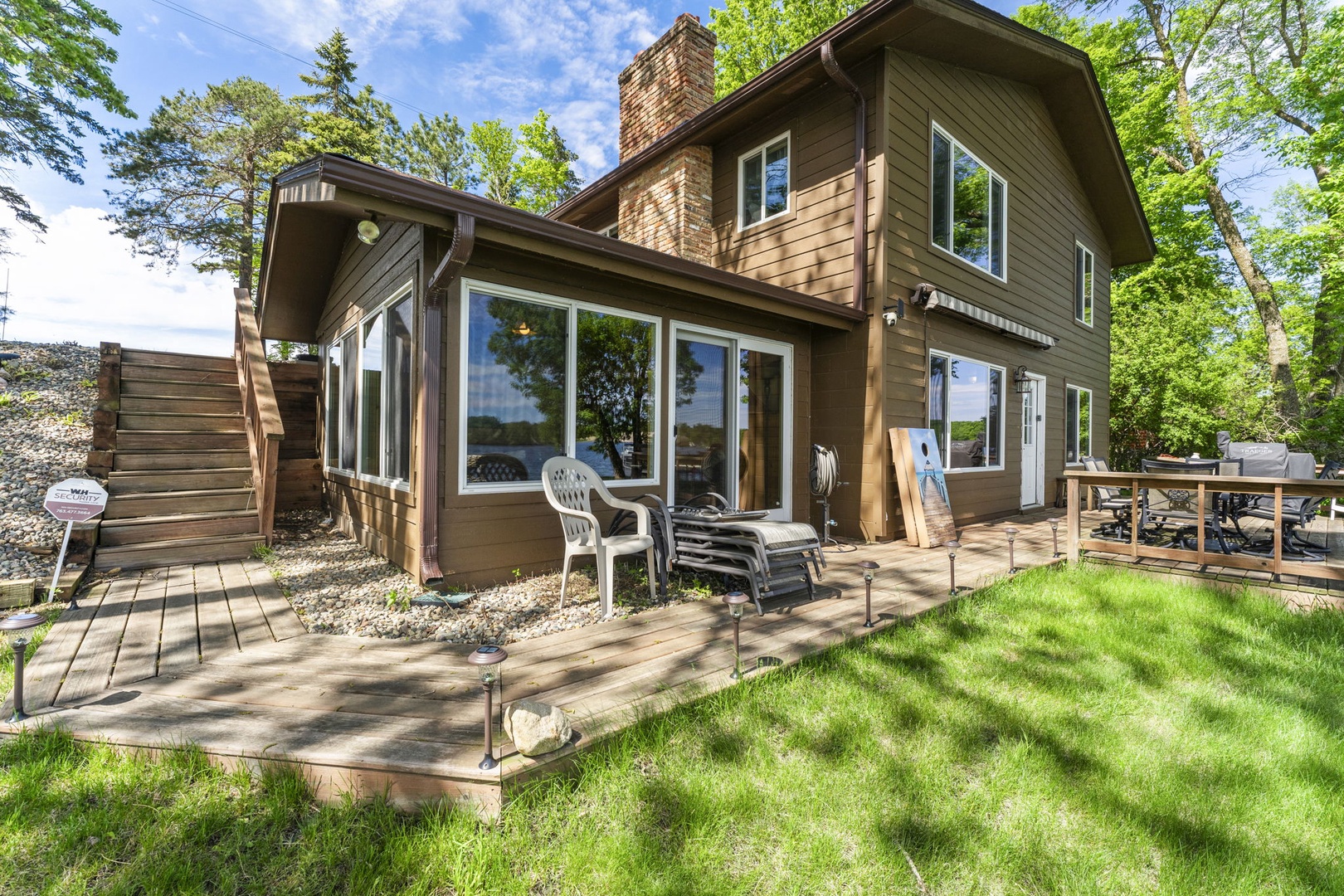 A charming two-story lakeside retreat featuring warm wood siding and a brick chimney, nestled among towering trees with a welcoming front lawn and outdoor seating for leisurely afternoons.