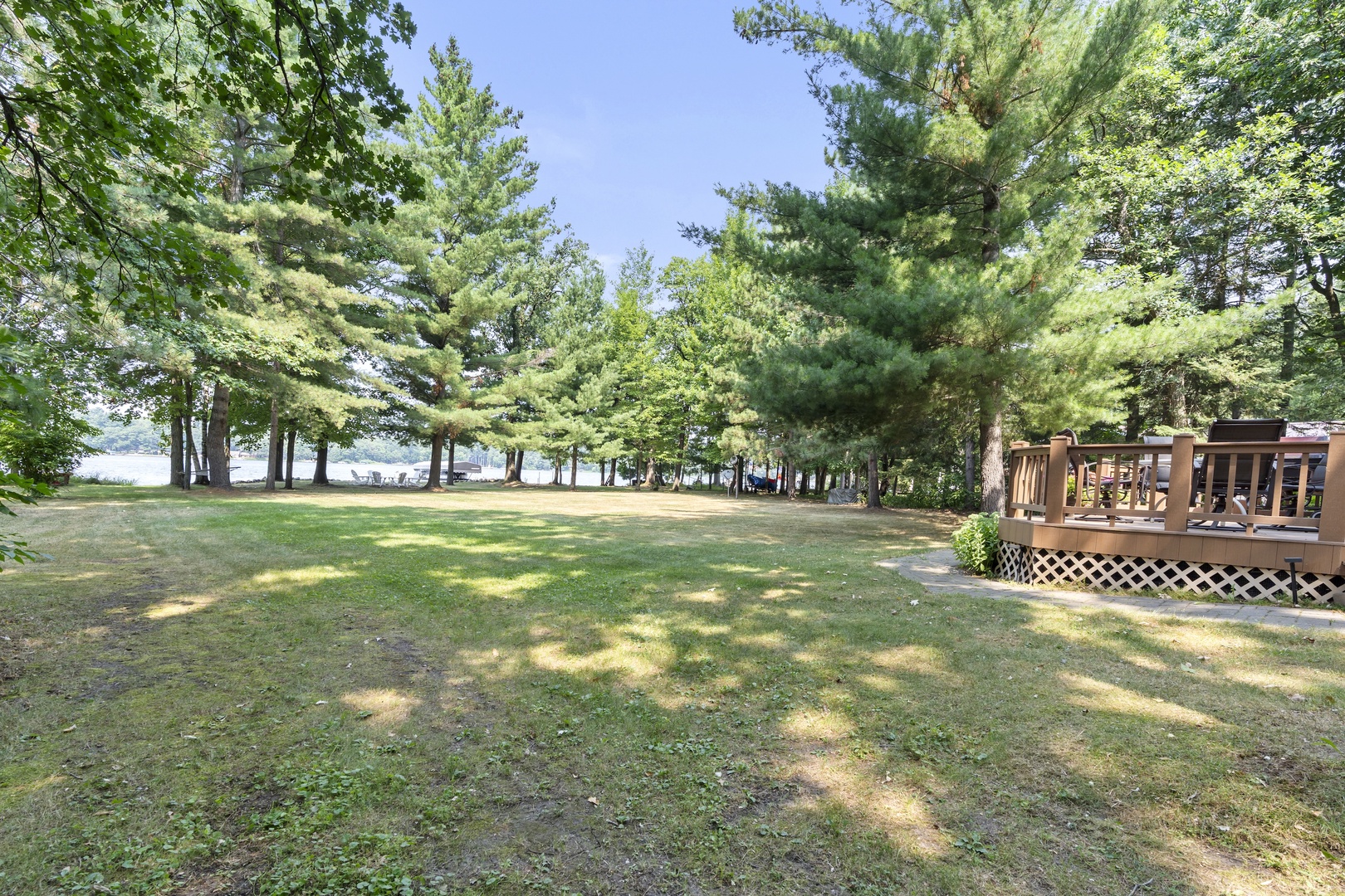The property sits on spacious grounds surrounded by mature trees, with a lake visible through the forest beyond. A wooden deck provides an ideal spot to enjoy the peaceful, natural setting.
