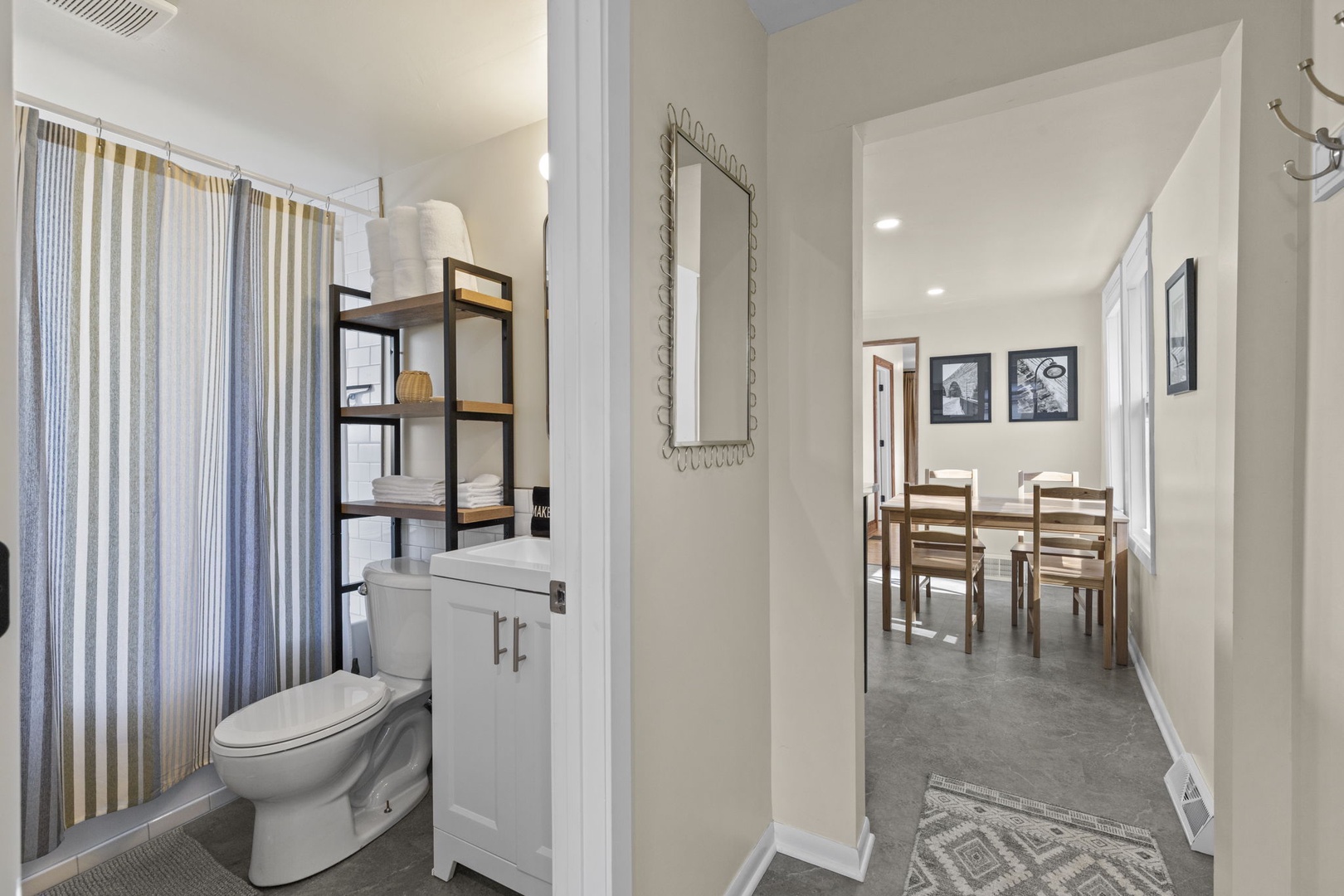 Full bathroom with tub/shower combo, striped curtain, open shelving with fresh towels, and white vanity — clean, bright, and well-stocked for your stay.