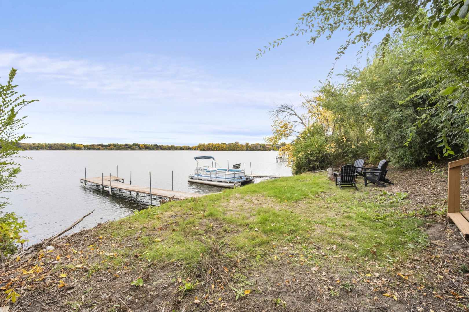 This tranquil lakefront setting features direct water access with a private dock and pontoon boat, framed by natural greenery and comfortable Adirondack chairs—perfect for peaceful mornings and sunset evenings by the lake.