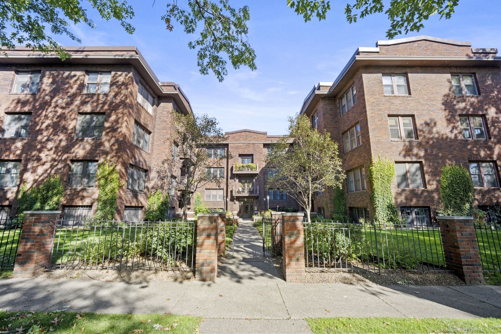 The property sits in a quiet residential neighborhood, framed by mature trees and well-maintained landscaping. Enter through the brick-pillared gate into a courtyard setting that welcomes you to the building.